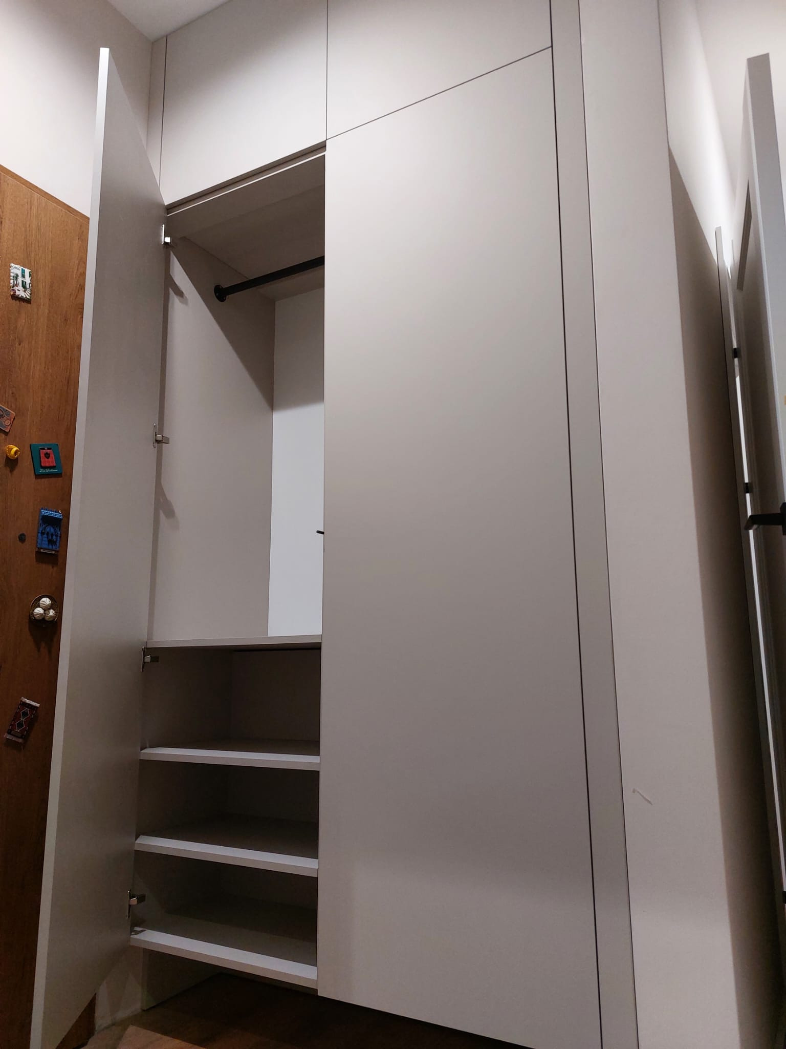 Jasna szafa wnękowa z otwartymi drzwiami, widoczne półki i drążek na ubrania. Minimalistyczny design, gładkie fronty, drewniane drzwi obok z dekoracjami.