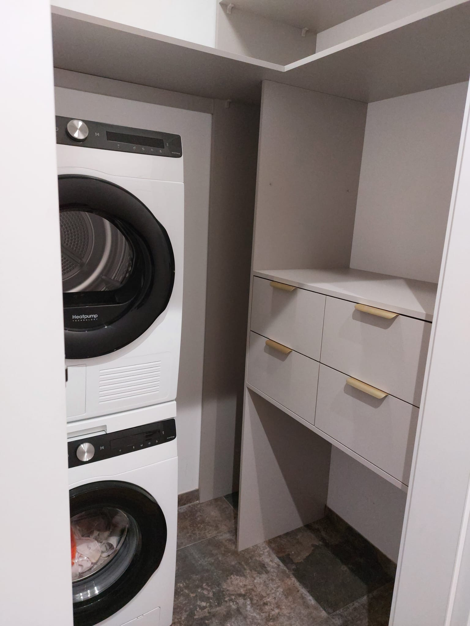 Biała pralka i suszarka w zabudowie szafy wnękowej z szufladami z uchwytami w kolorze złota. Minimalistyczny design, szara kolorystyka, oświetlenie górne.