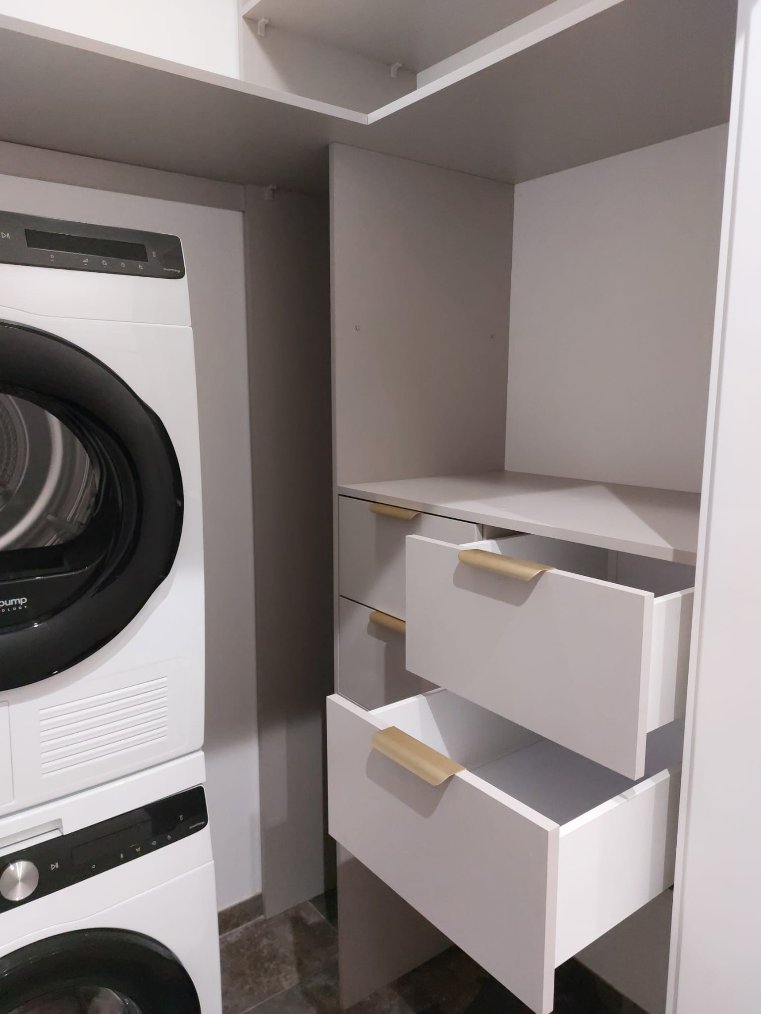 Biała szafa wnękowa z szufladami z uchwytami w kolorze złotym, obok pralka i suszarka w zabudowie. Minimalistyczny design, szare ściany, ciemna podłoga.
