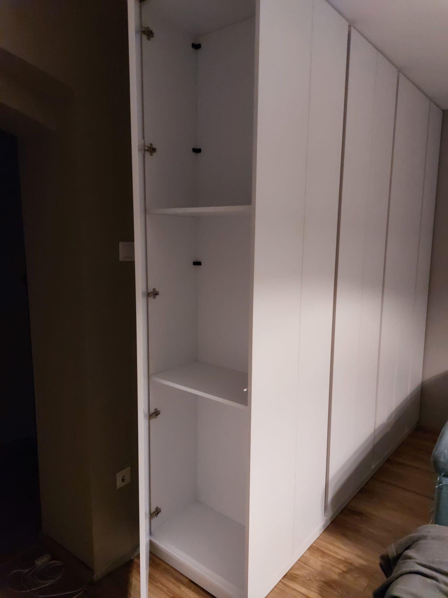 Biała szafa wnękowa z otwartymi drzwiami, widoczne półki wewnątrz. Minimalistyczny design, jasne wnętrze, ciemne tło ściany. Ujęcie z bliska, ukazujące detale wykończenia.