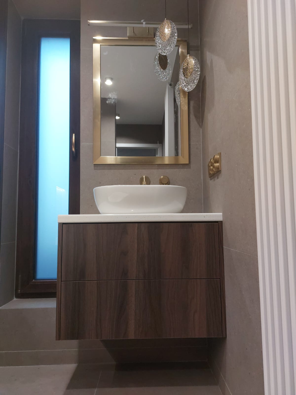 Nowoczesna łazienka z drewnianą szafką pod umywalką, białym blatem i owalną umywalką. Lustro w złotej ramie i designerskie lampy wiszące dodają elegancji.