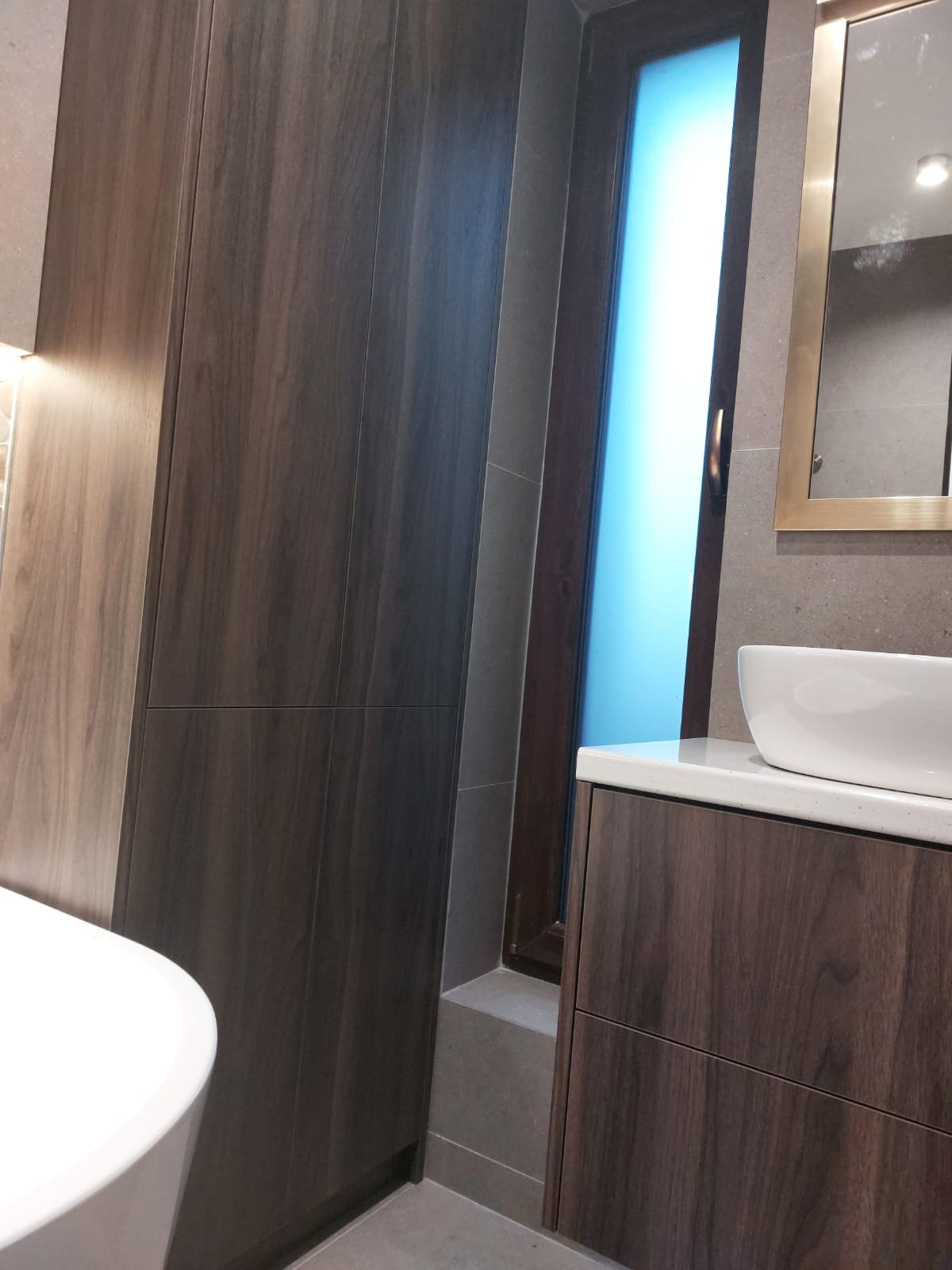 Nowoczesna łazienka z szafkami z ciemnego drewna, białą umywalką nablatową i lustrem w złotej ramie. Minimalistyczny design, stonowana kolorystyka, eleganckie wykończenie.