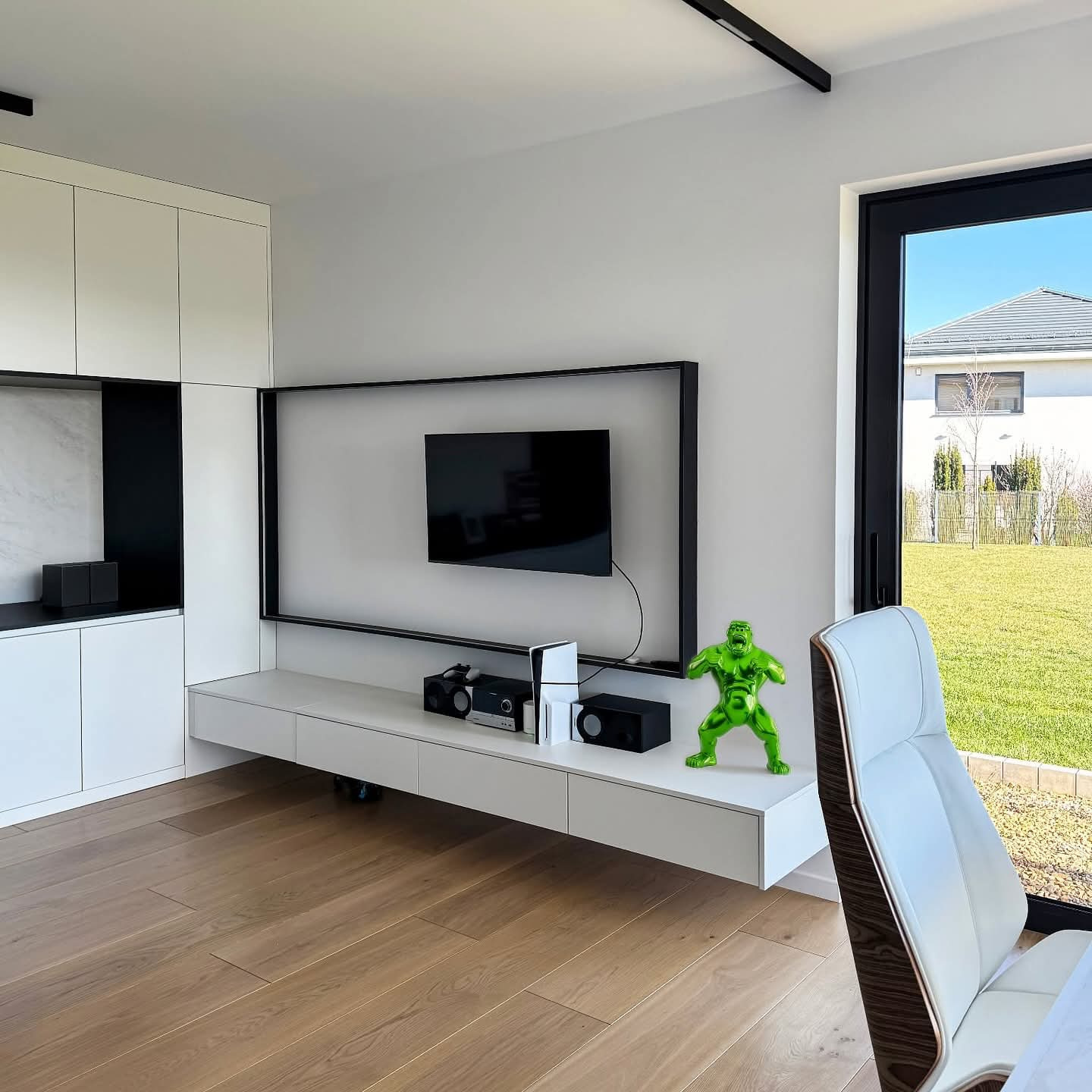 Nowoczesny salon z białymi meblami na wymiar, drewnianą podłogą i zieloną figurką Hulka. Minimalistyczny design z czarnymi akcentami i dużym oknem na ogród.