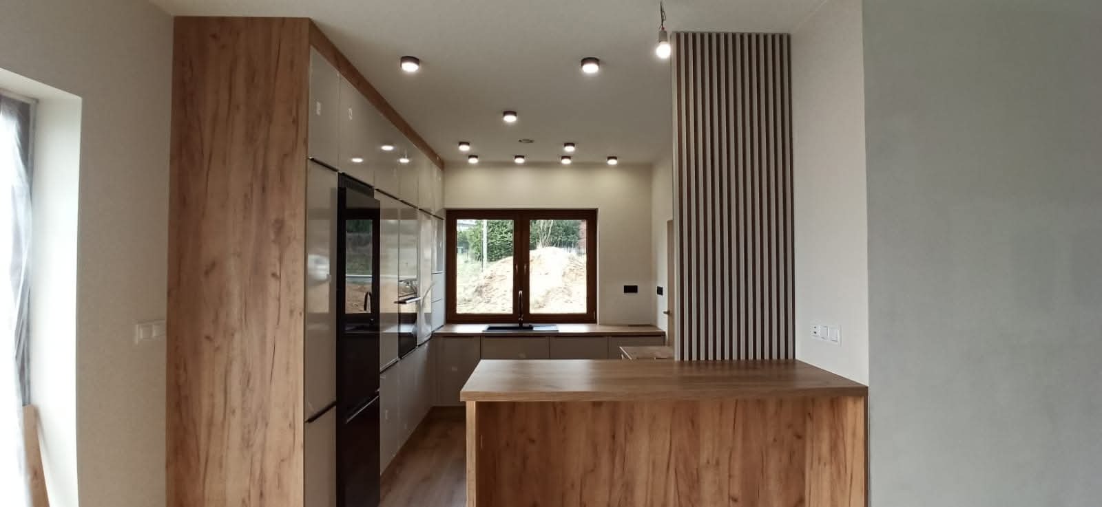 Nowoczesna kuchnia z drewnianymi akcentami: wysoka zabudowa, wyspa z blatem o strukturze drewna i dekoracyjna ścianka z pionowych listew. Jasne wnętrze z oknem.