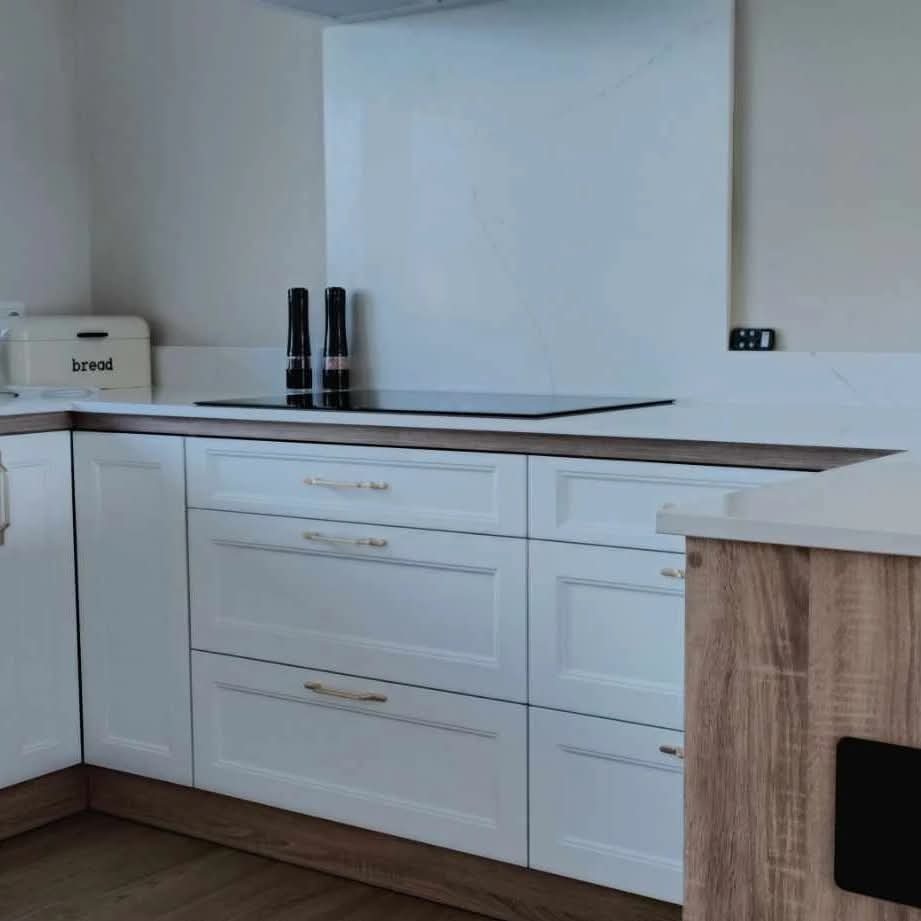 Białe szafki kuchenne z drewnianym blatem i złotymi uchwytami, uzupełnione o płytę indukcyjną i akcesoria. Widoczny fragment wyspy kuchennej z drewnianym wykończeniem.