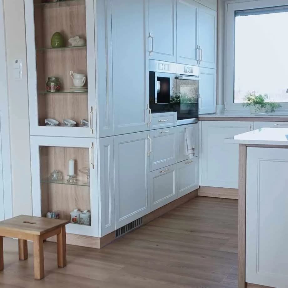 Jasna kuchnia z przeszklonymi szafkami, drewnianymi akcentami i małym stołkiem. Widoczne AGD w zabudowie i drewniana podłoga.