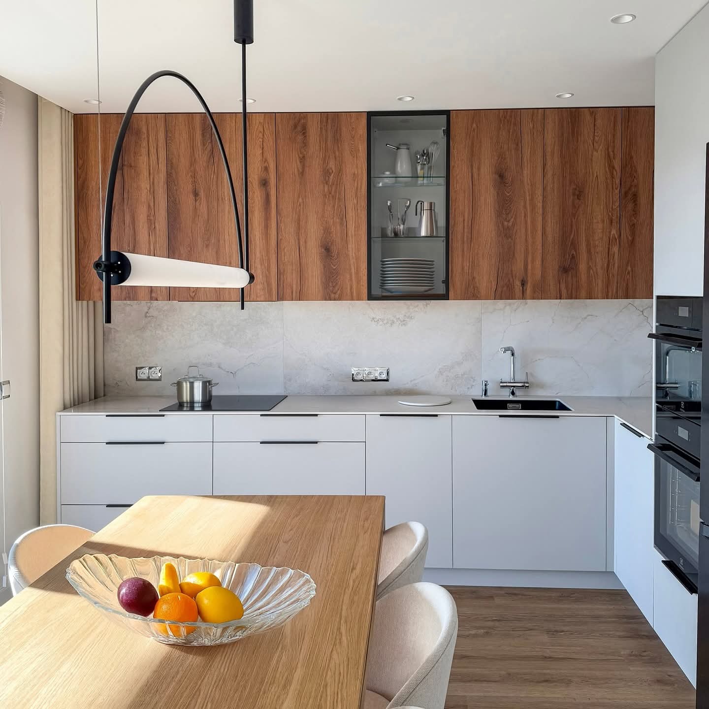 Nowoczesna kuchnia z drewnianymi szafkami górnymi i białymi dolnymi. Na pierwszym planie stół z owocami w szklanej misie. Minimalistyczna lampa nad stołem.