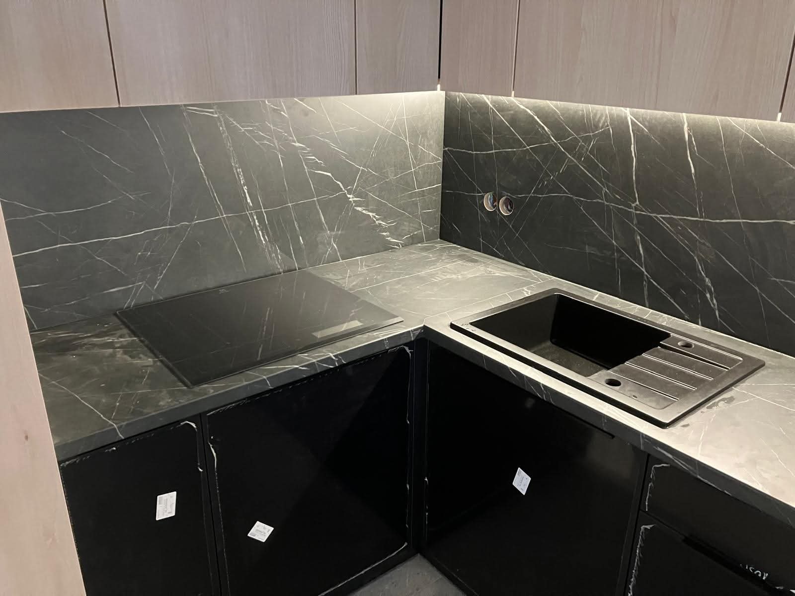 Ciemna kuchnia z marmurowym blatem i płytą indukcyjną. Czarna zabudowa szafek, nowoczesny zlew. Oświetlenie LED pod szafkami górnymi. Minimalistyczny design.