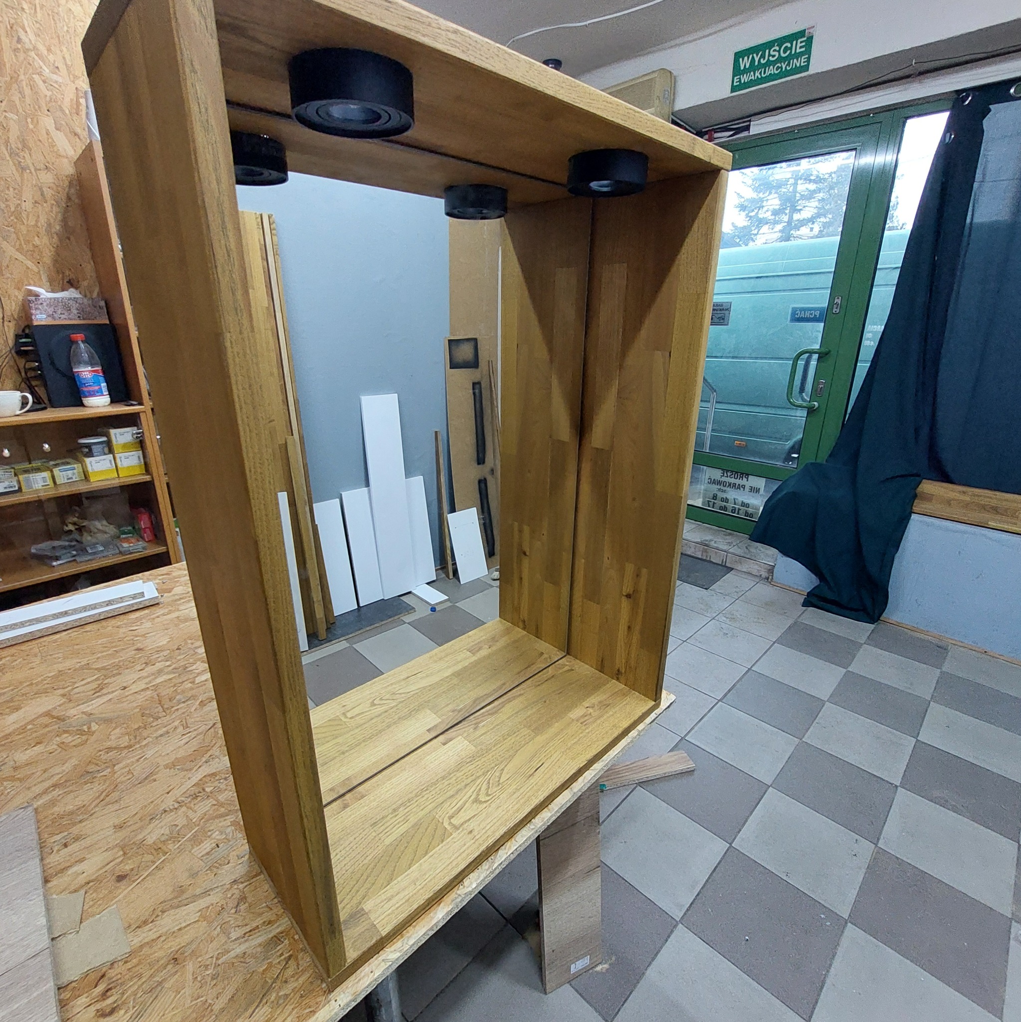 Surowa, drewniana rama meblowa z oświetleniem punktowym, w warsztacie. Widoczne elementy konstrukcyjne i tło z materiałami stolarskimi. Ujęcie z bliska, ukazujące detale.