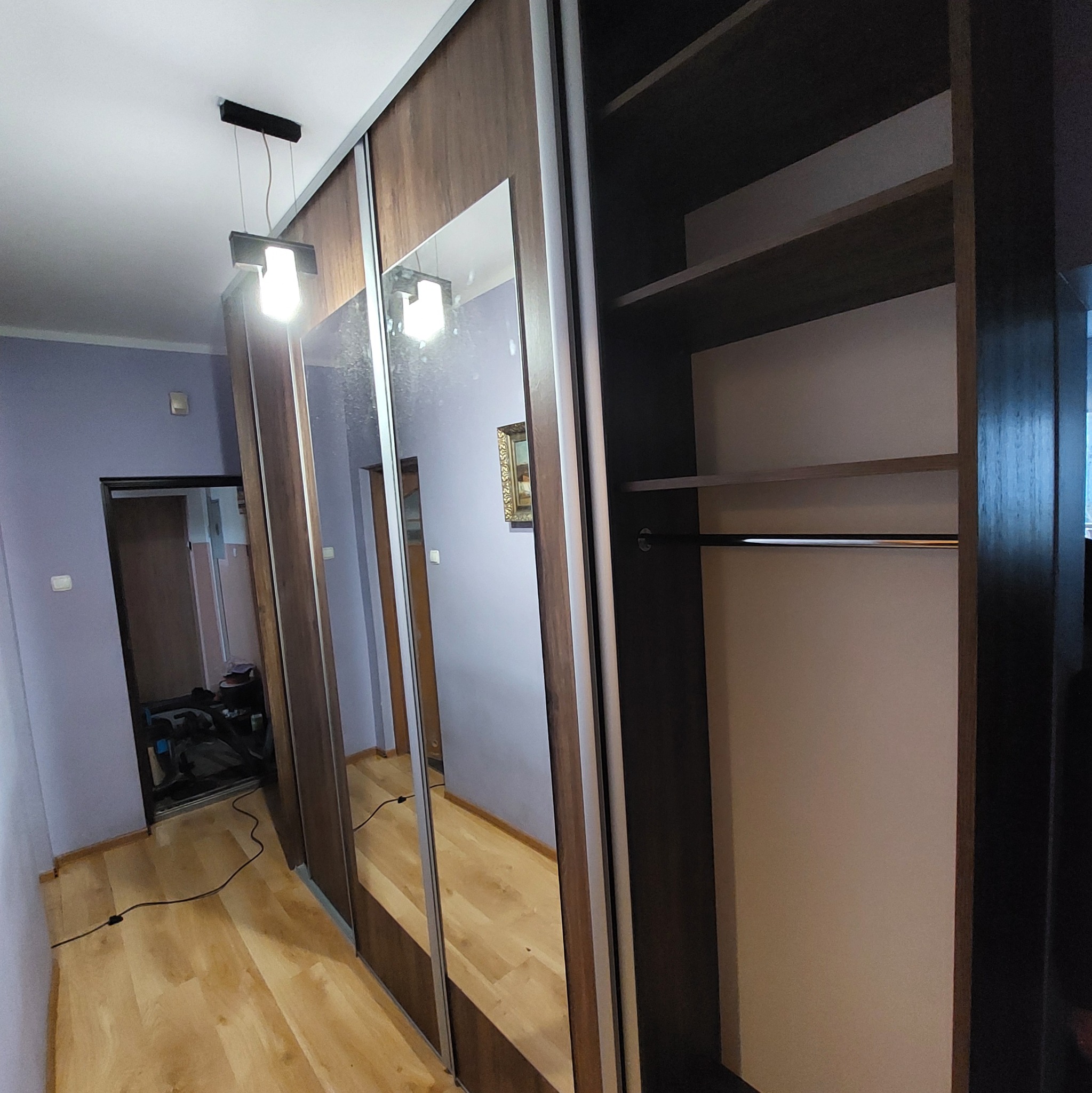 Wnętrze z szafą wnękową z przesuwnymi drzwiami lustrzanymi, oświetleniem sufitowym i drewnianą podłogą. Widoczne półki i drążek na ubrania.