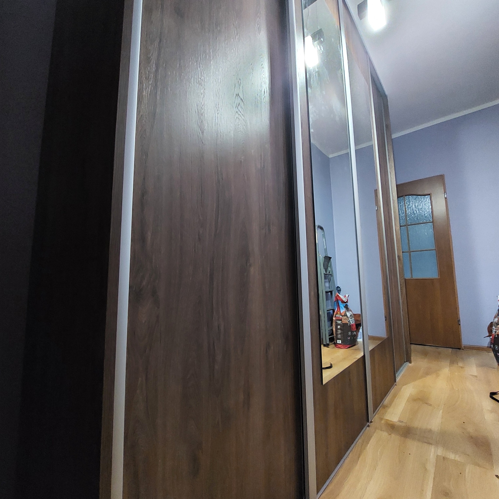 Szafa przesuwna z lustrami w Radlinie. Brązowe fronty z widoczną strukturą drewna, srebrne obramowania. Widoczne odbicie wnętrza pokoju i drzwi.