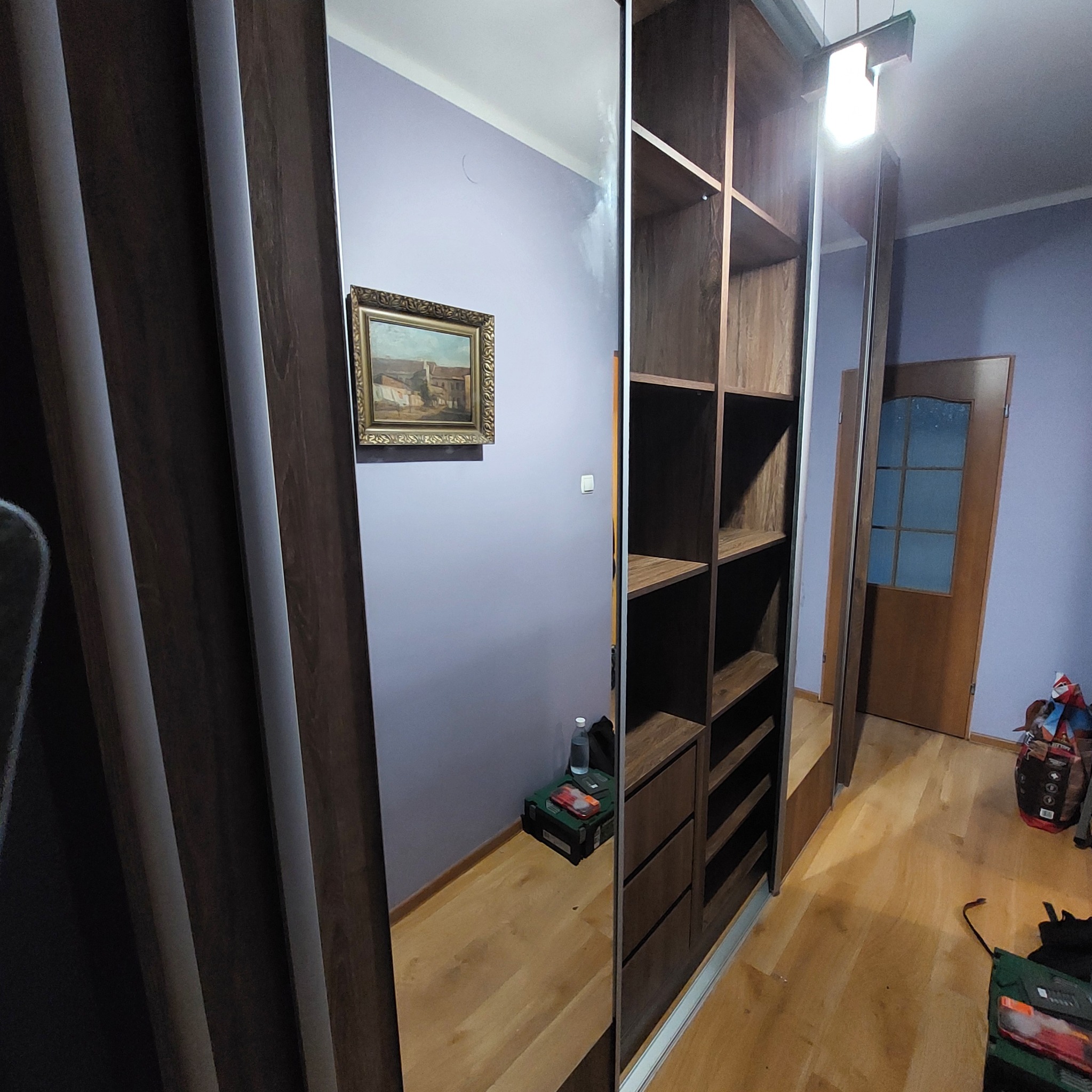 Szafa wnękowa z lustrzanymi drzwiami przesuwnymi, z otwartymi półkami i szufladami, w pokoju z obrazem na ścianie i drewnianą podłogą.