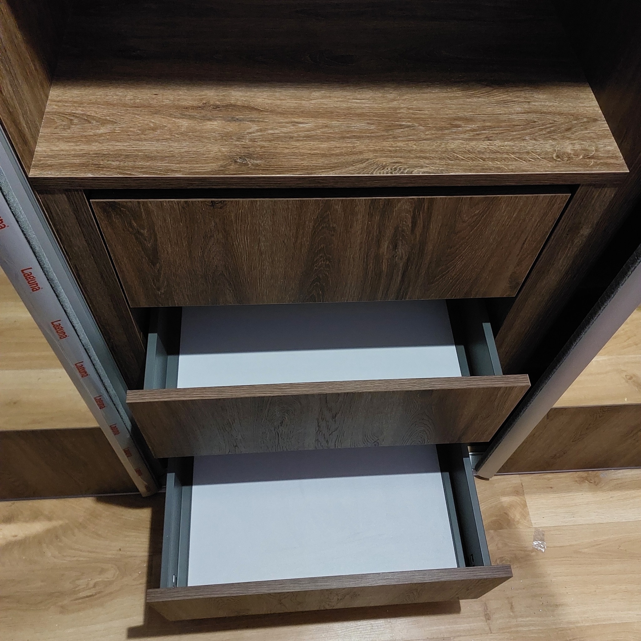 Szafa z okleiną drewnopodobną, z trzema wysuniętymi szufladami z białym dnem. Widoczne prowadnice szuflad. Ujęcie z bliska, ukazujące detale wykończenia.