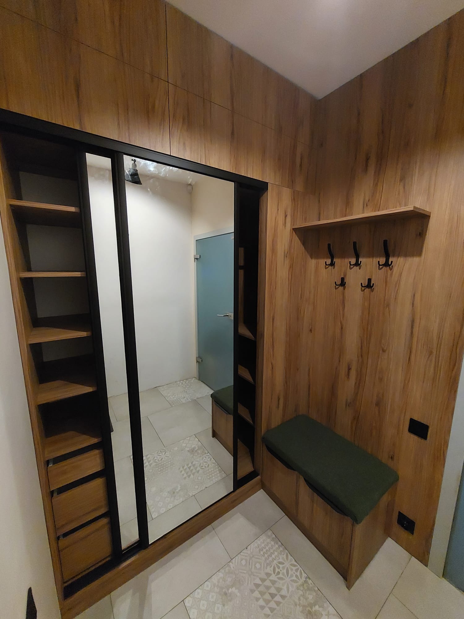 Wnętrze przedpokoju z zabudową: szafa przesuwna z lustrem, drewniana boazeria, półka z wieszakami i siedzisko z zieloną poduszką. Całość utrzymana w ciepłej tonacji drewna.