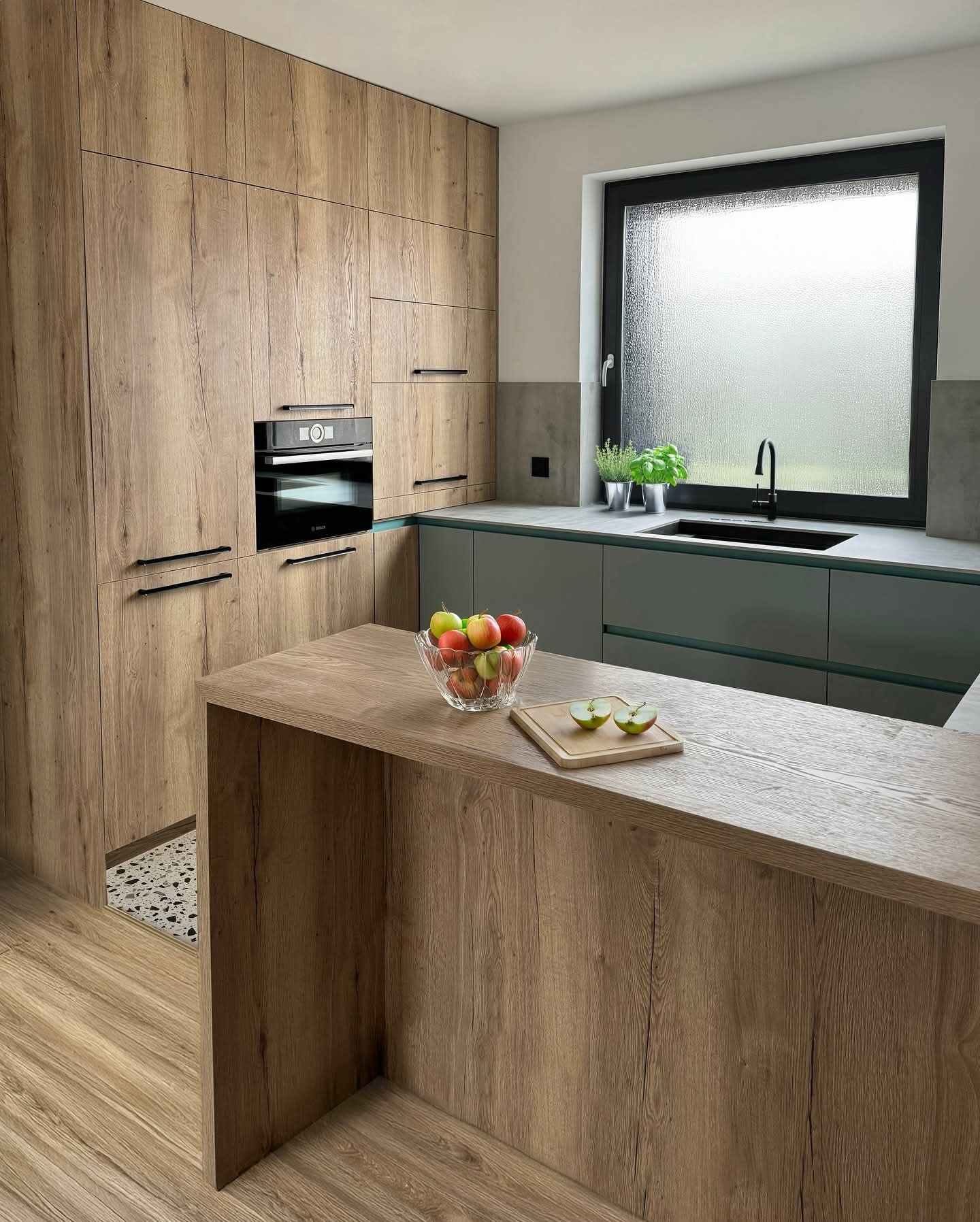 Nowoczesna kuchnia z drewnianymi frontami, czarnymi uchwytami i oknem z widokiem na zewnątrz. Na blacie szklana misa z jabłkami i deska z przekrojonym jabłkiem.