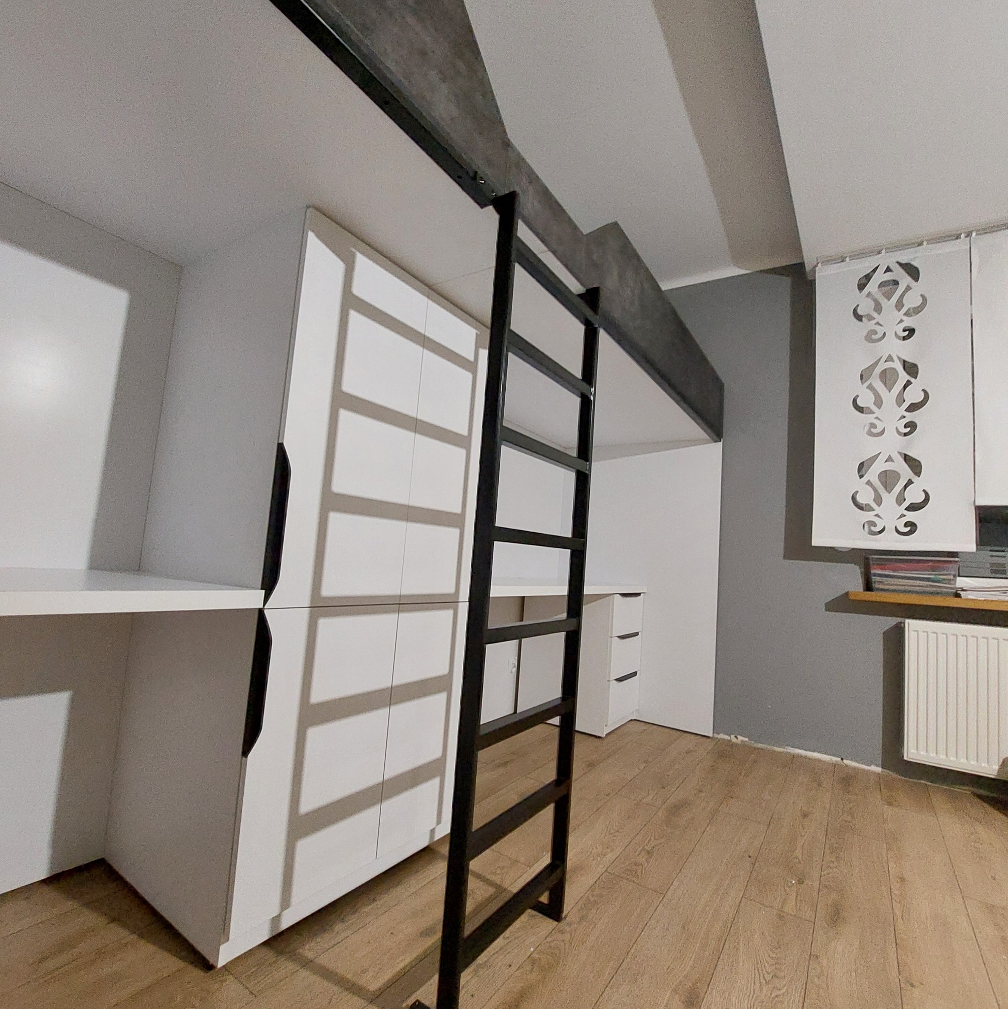 Biała zabudowa meblowa z biurkiem i szafą, połączona z antresolą. Czarna drabina prowadzi na górę. Nowoczesny design, jasne wnętrze z drewnianą podłogą.