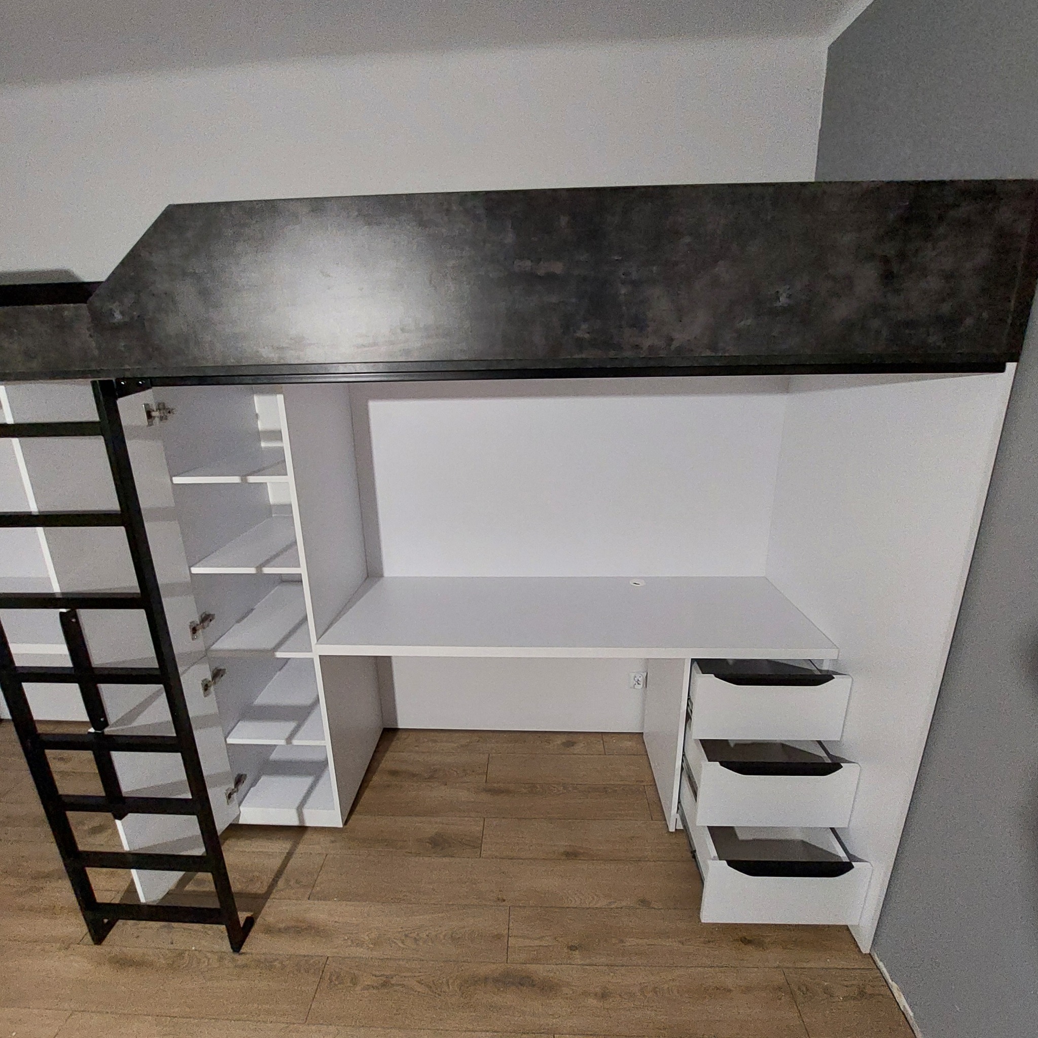 Funkcjonalne łóżko piętrowe z biurkiem i szafkami w zabudowie, czarno-biała kolorystyka, metalowa drabinka, drewniana podłoga. Nowoczesny design.