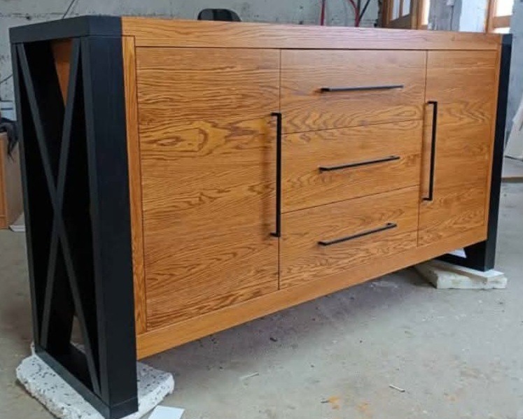 Drewniana komoda z czarnymi elementami, widoczne szafki i szuflady z podłużnymi uchwytami. Wykończenie w ciepłym odcieniu drewna z wyraźnym usłojeniem.