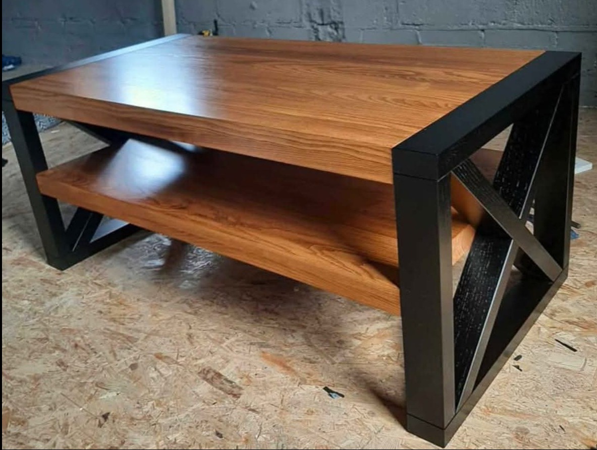 Stolik kawowy z blatem z drewna o ciepłym odcieniu i czarną, geometryczną podstawą. Widoczna półka pod blatem, całość na tle płyty OSB.