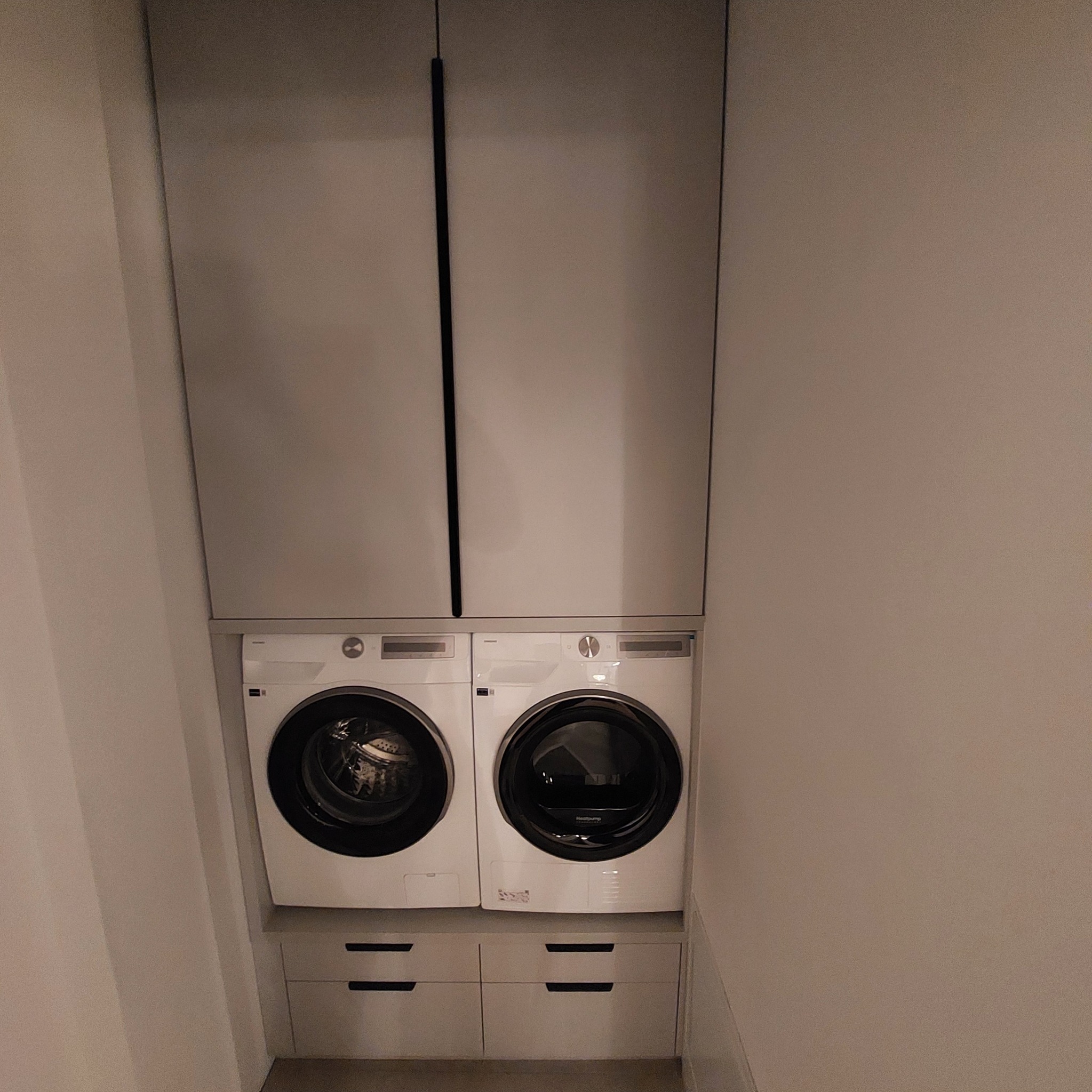 Szara zabudowa meblowa z dwiema pralkami i szafkami wiszącymi. Minimalistyczny design, symetryczny układ, nowoczesne wykończenie. Widok z przodu, oświetlenie naturalne.