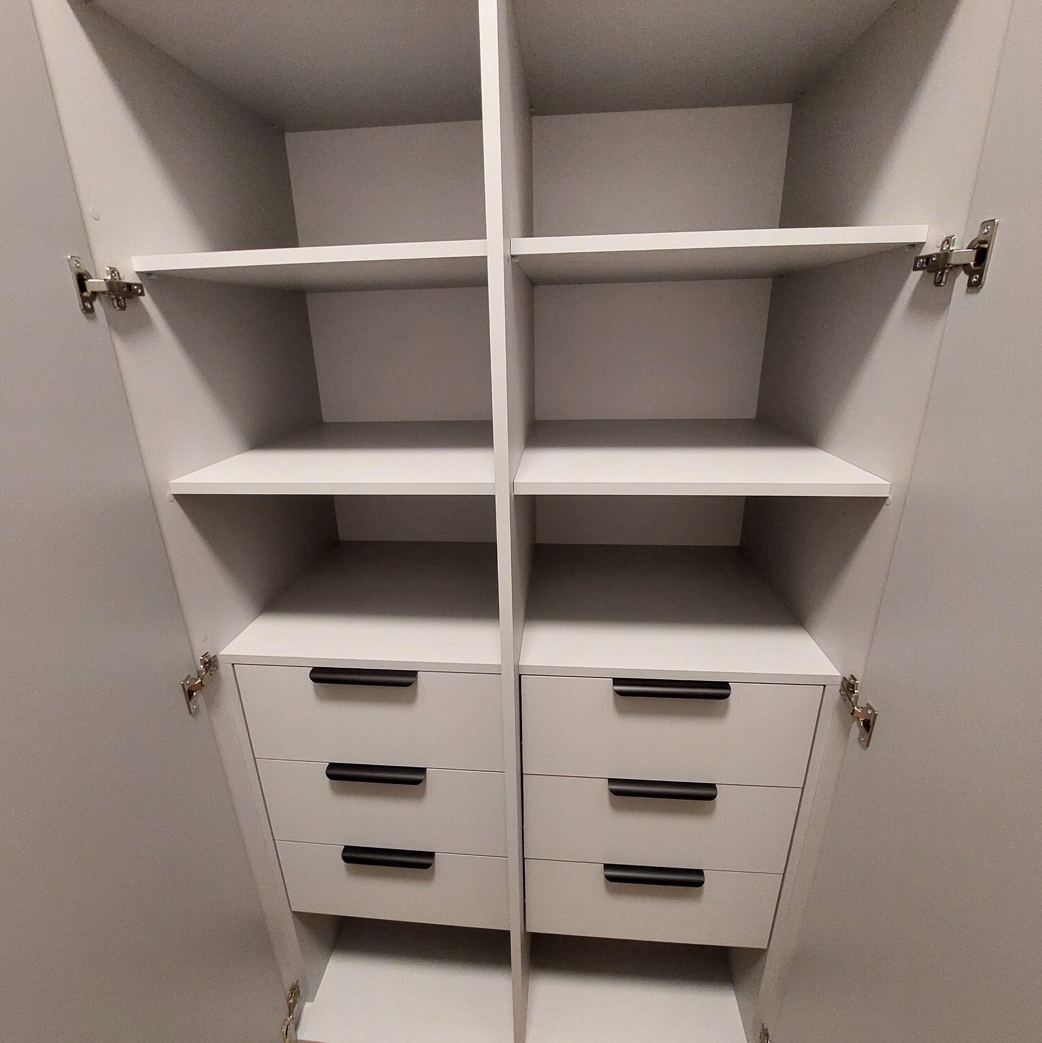 Wnętrze szafy z półkami i szufladami w odcieniach szarości, widoczne zawiasy i czarne uchwyty. Minimalistyczny design, symetryczny układ.