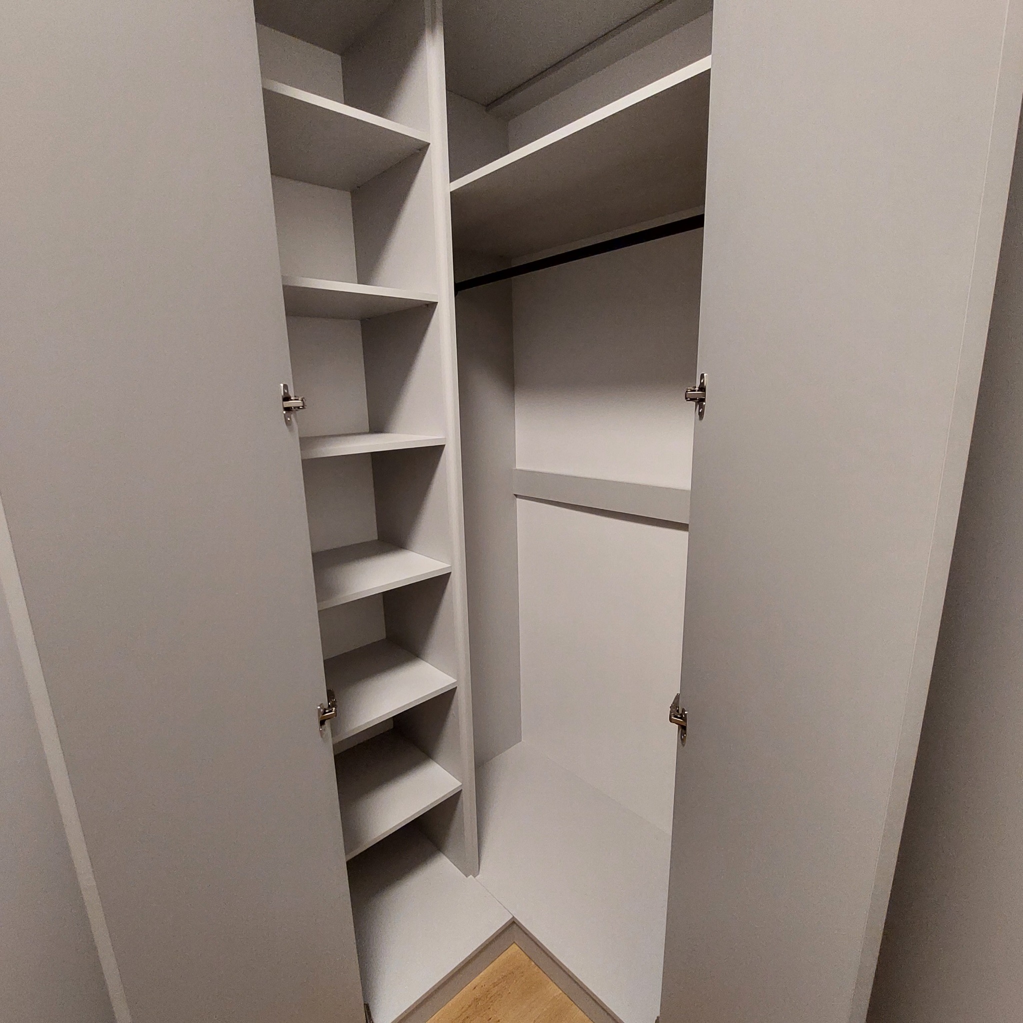 Otwarta, narożna szafa wnękowa z półkami po lewej i drążkiem na ubrania po prawej stronie. Jasnoszary kolor, minimalistyczny design, widoczny fragment podłogi.