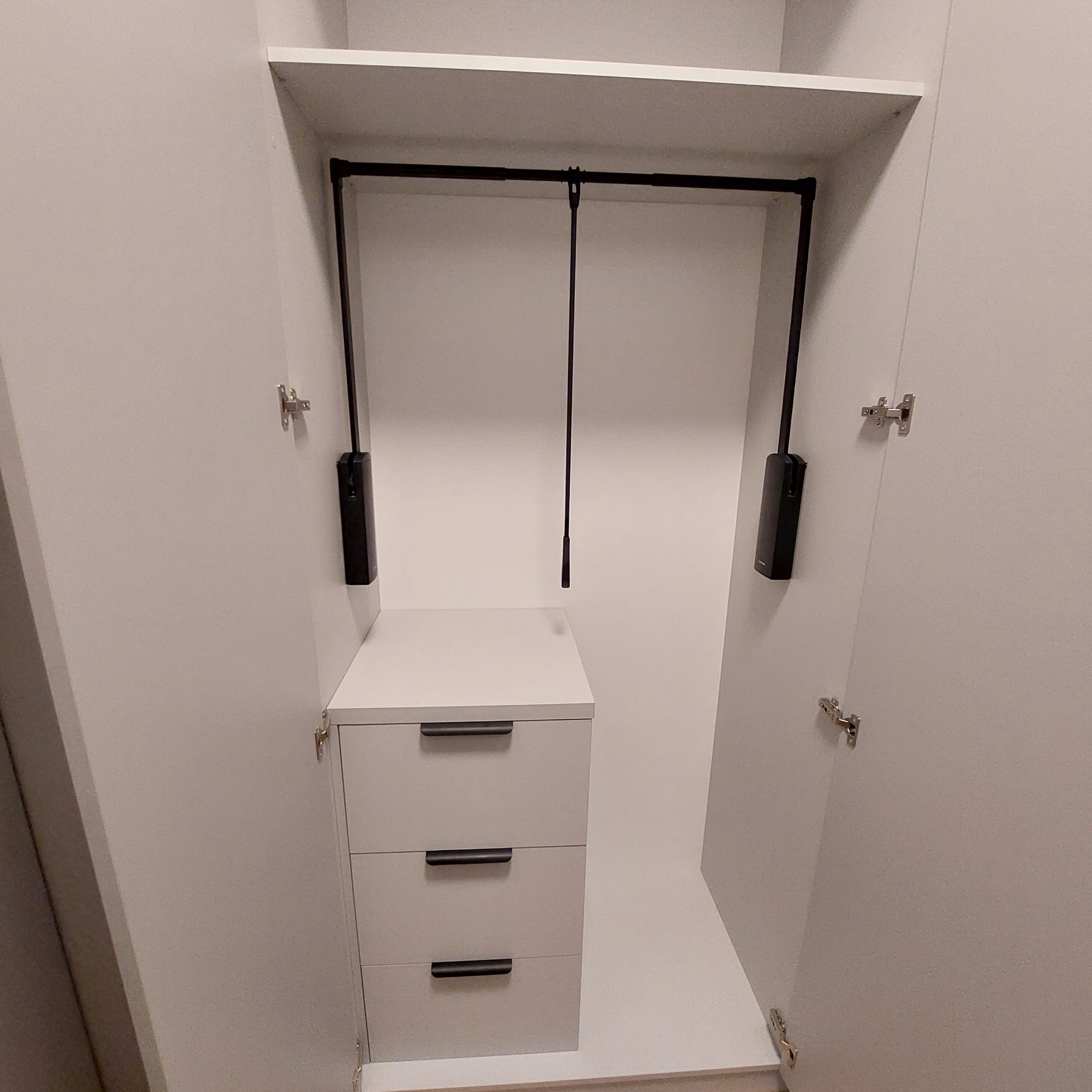 Wnętrze szafy wnękowej z trzema szufladami i drążkiem na ubrania z systemem opuszczania. Białe fronty, minimalistyczne uchwyty, otwarte drzwi.