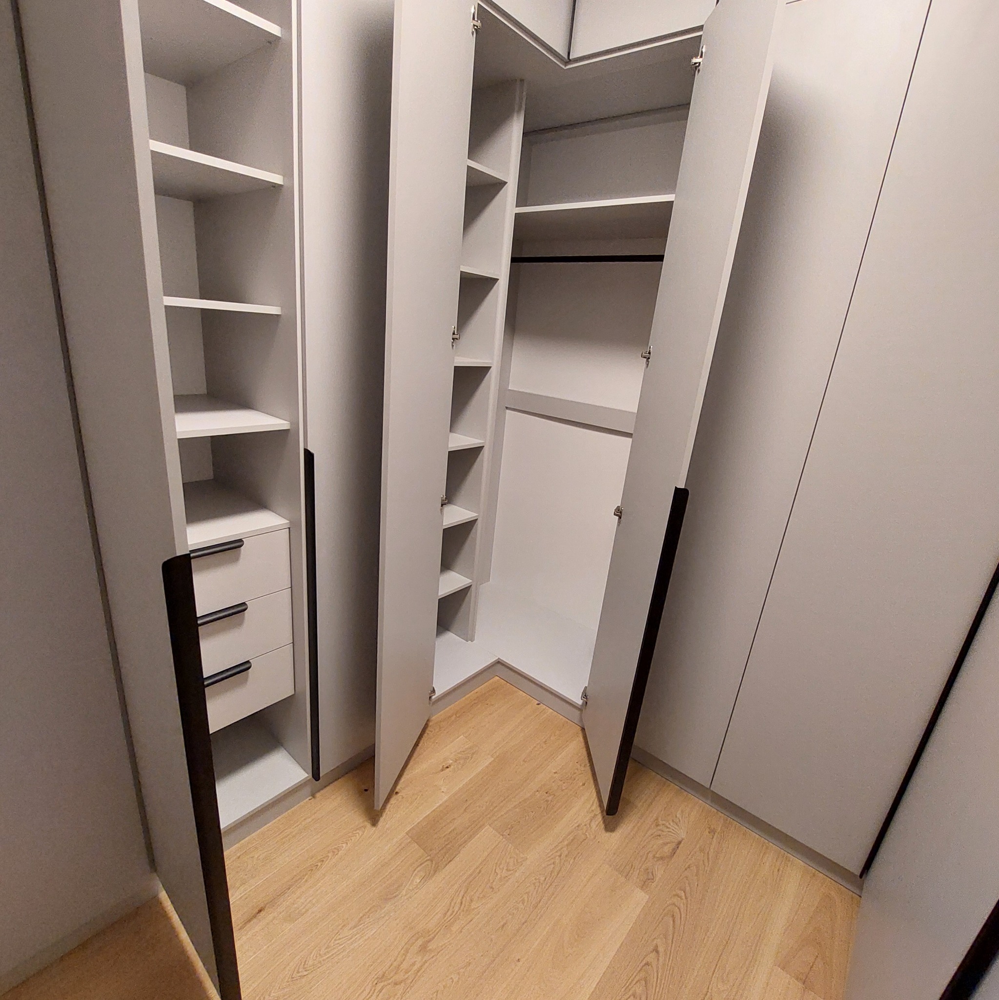 Jasnoszara szafa narożna z otwartymi drzwiami, widoczne półki i szuflady z czarnymi uchwytami, na podłodze panele w odcieniach drewna.