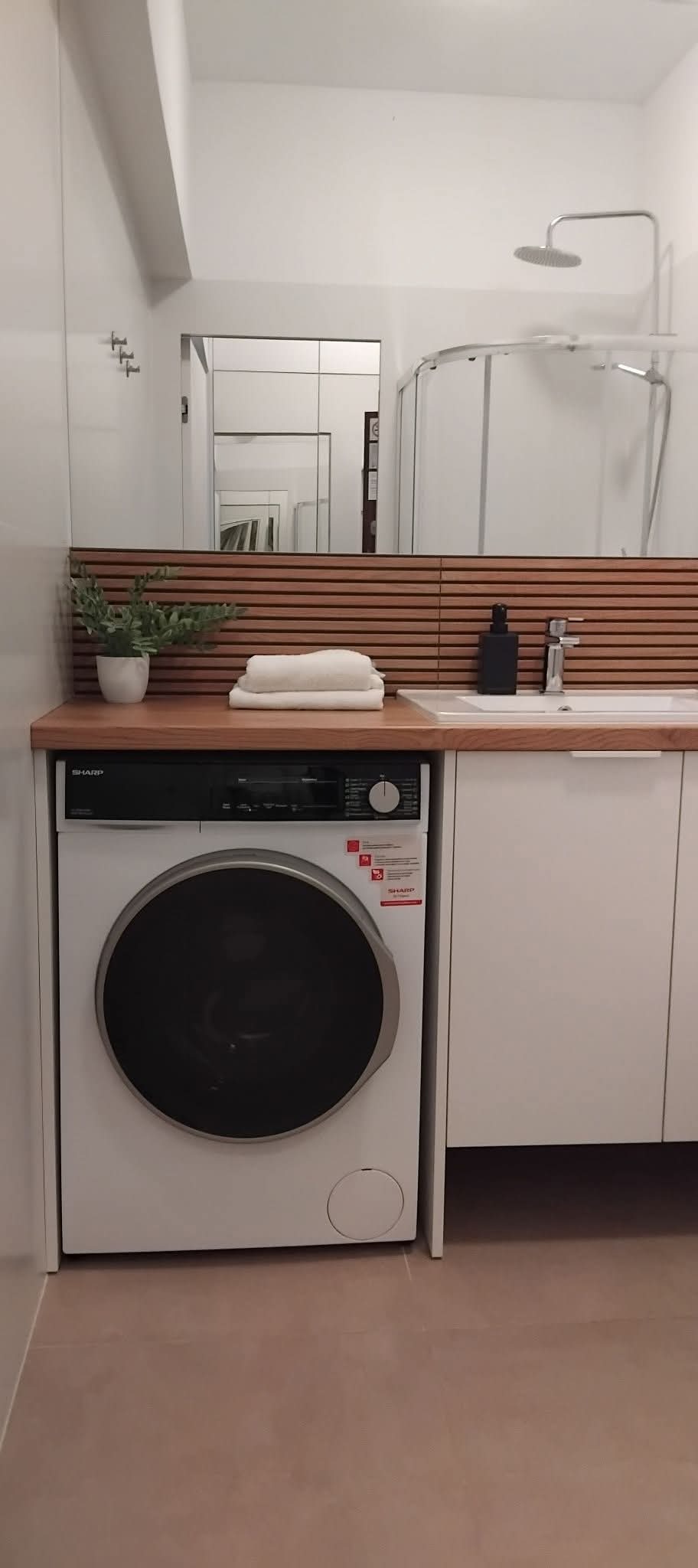 Nowoczesna łazienka z pralką Sharp, drewnianym panelem dekoracyjnym, białymi ręcznikami i minimalistyczną armaturą. Widoczna kabina prysznicowa w tle.