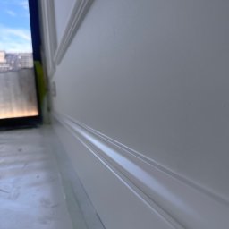 NJmalers - Świeżo pomalowana na biało ściana z ozdobną listwą przypodłogową i zabezpieczoną folią podłogą, widok z dołu, fragment okna z widokiem na niebo.