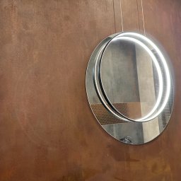 NJmalers - Rdzawo-brązowa ściana z włącznikiem i okrągłym lustrem LED, odbijającym fragment łazienki z mozaiką i umywalką. Dekoracyjne wykończenie ściany.