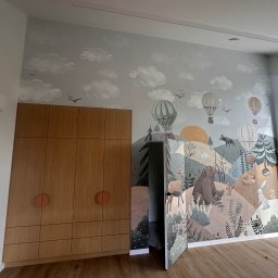 NJmalers - Pokój dziecięcy po malowaniu: ściana z motywem lasu i zwierząt, obok drewniana szafa z okrągłymi uchwytami. Widoczne okno i drewniana podłoga.