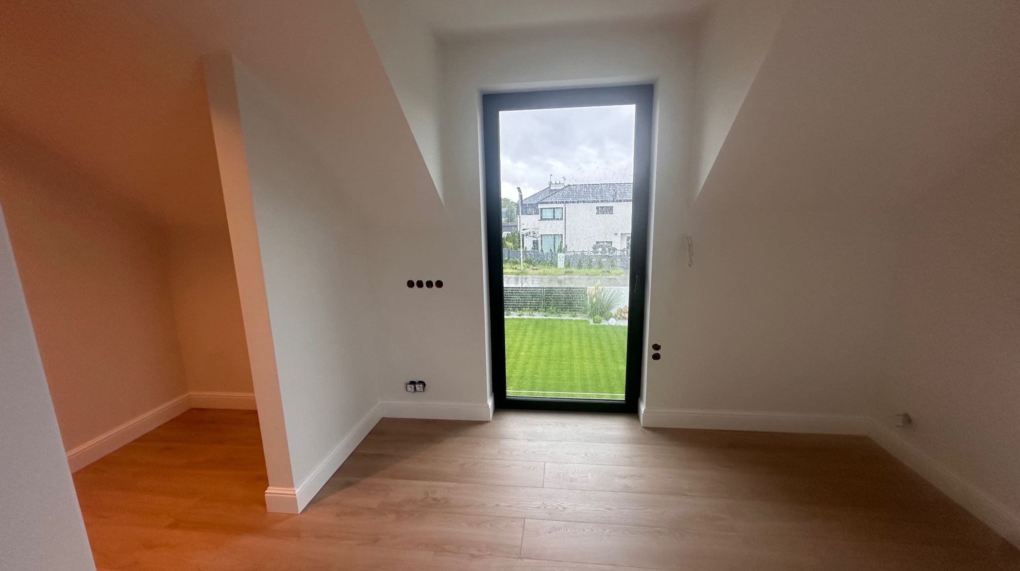 Jasne, świeżo wykończone poddasze w Szczecinie z drewnianą podłogą i oknem z widokiem na ogród. Widoczne gniazdka elektryczne i otwory na kable.