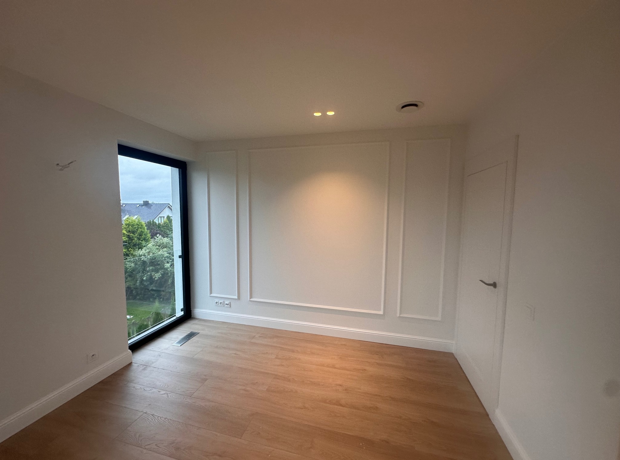 Jasne wnętrze po remoncie z drewnianą podłogą, białymi ścianami z dekoracyjnymi listwami i dużym oknem z widokiem na zieleń. Widoczne gniazdka elektryczne.