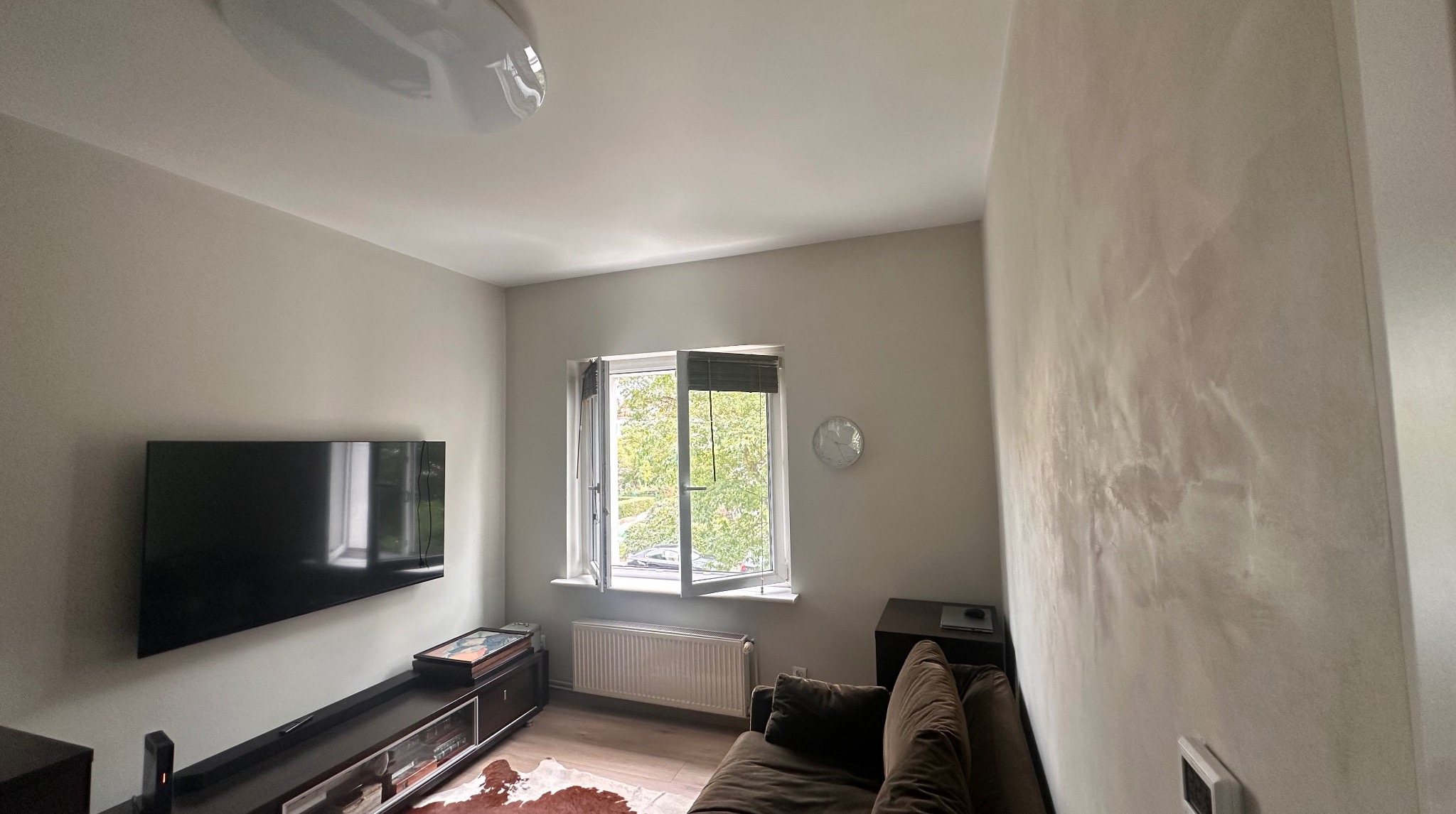 Jasne wnętrze po remoncie: ściany w odcieniach szarości z dekoracyjną fakturą, okno z żaluzjami, telewizor na ścianie. Widok na salon z elementami wyposażenia.