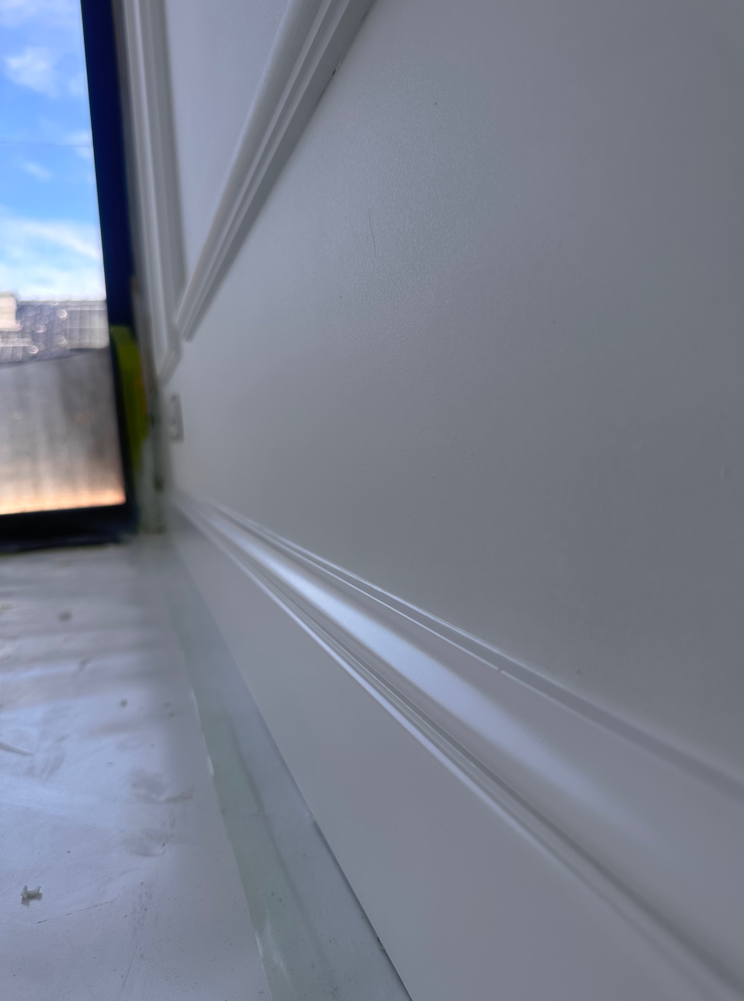 Świeżo pomalowana na biało ściana z ozdobną listwą przypodłogową i zabezpieczoną folią podłogą, widok z dołu, fragment okna z widokiem na niebo.