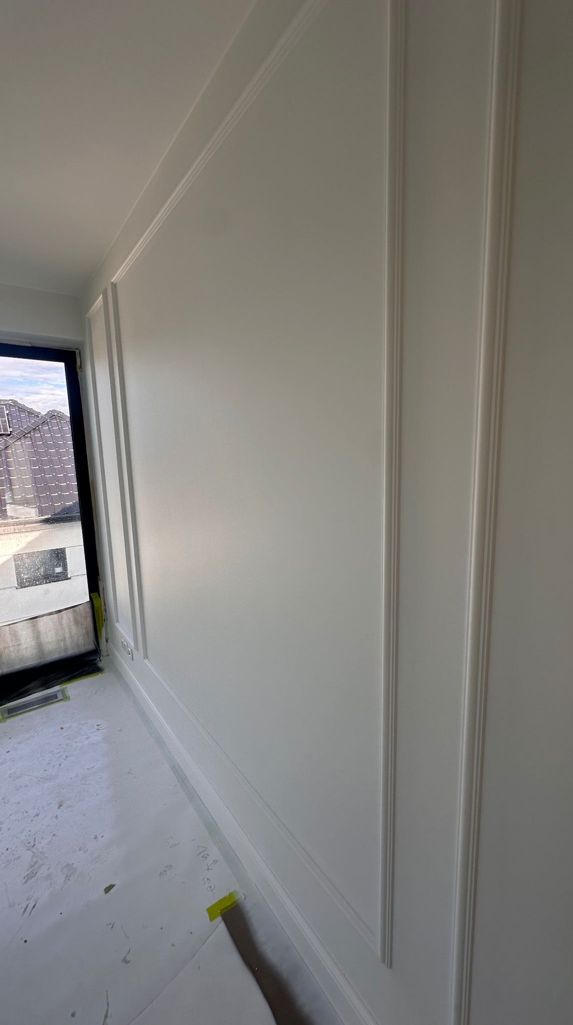 Biała ściana z dekoracyjnymi listwami w jasnym wnętrzu, widok z boku, okno z widokiem na dachy w tle. Podłoga zabezpieczona folią malarską.