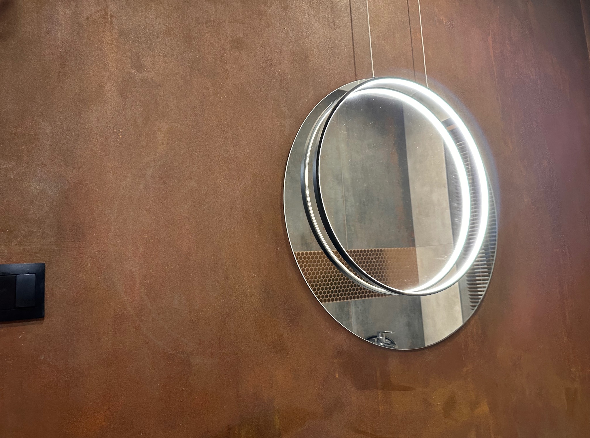 Rdzawo-brązowa ściana z włącznikiem i okrągłym lustrem LED, odbijającym fragment łazienki z mozaiką i umywalką. Dekoracyjne wykończenie ściany.