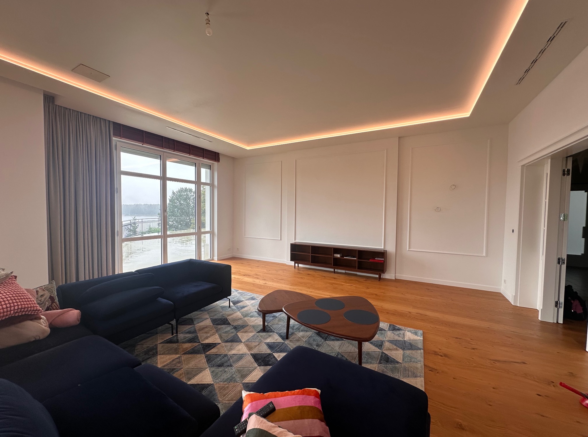 Jasne wnętrze salonu z oknem na jezioro, oświetleniem LED na suficie i ciemnoniebieską sofą na wzorzystym dywanie. Minimalistyczny wystrój, drewniana podłoga.