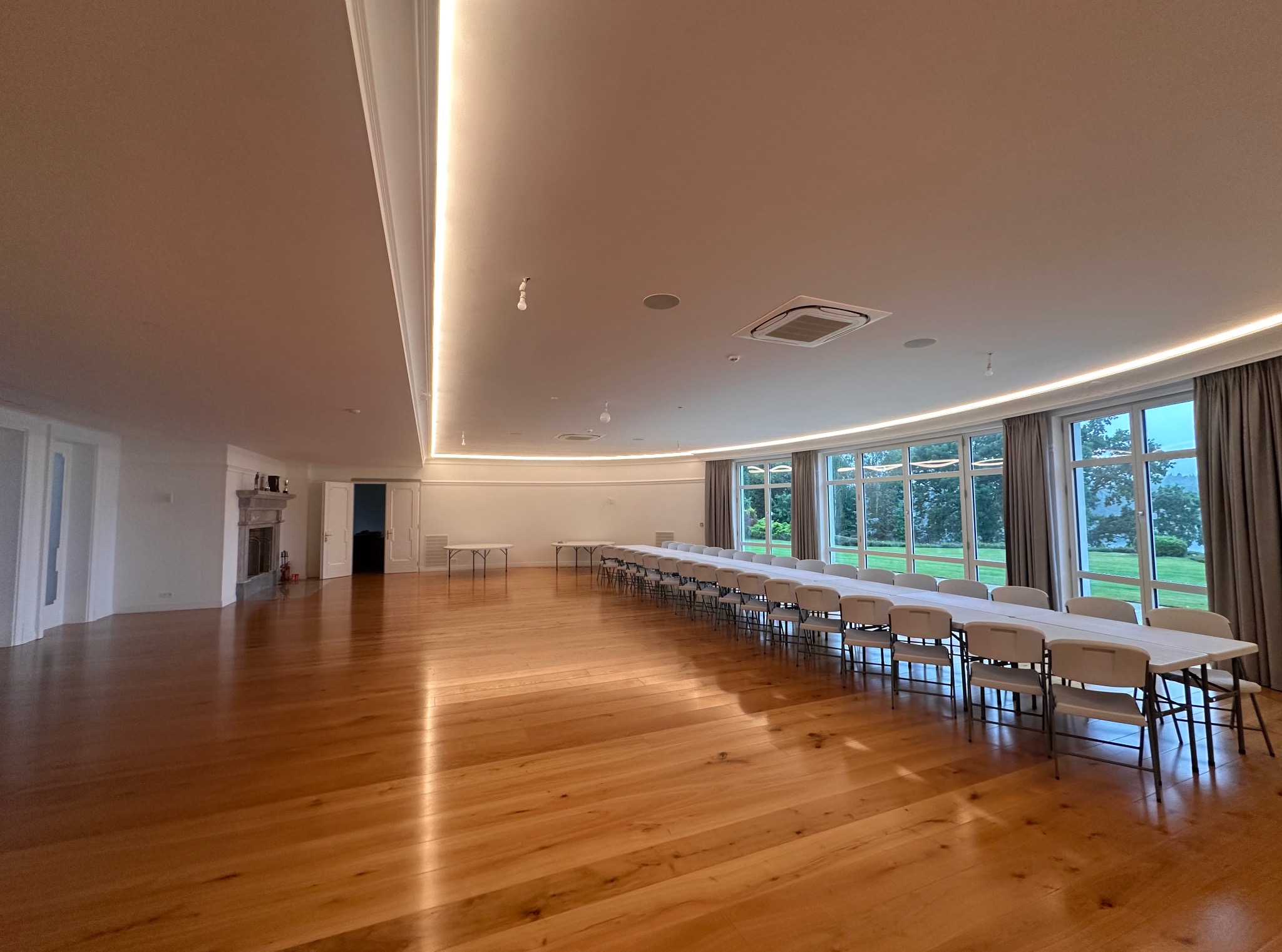 Jasne, przestronne wnętrze z drewnianą podłogą, długim stołem i rzędem krzeseł pod oknami. Sufit z oświetleniem LED. Widok na zieleń za oknem.