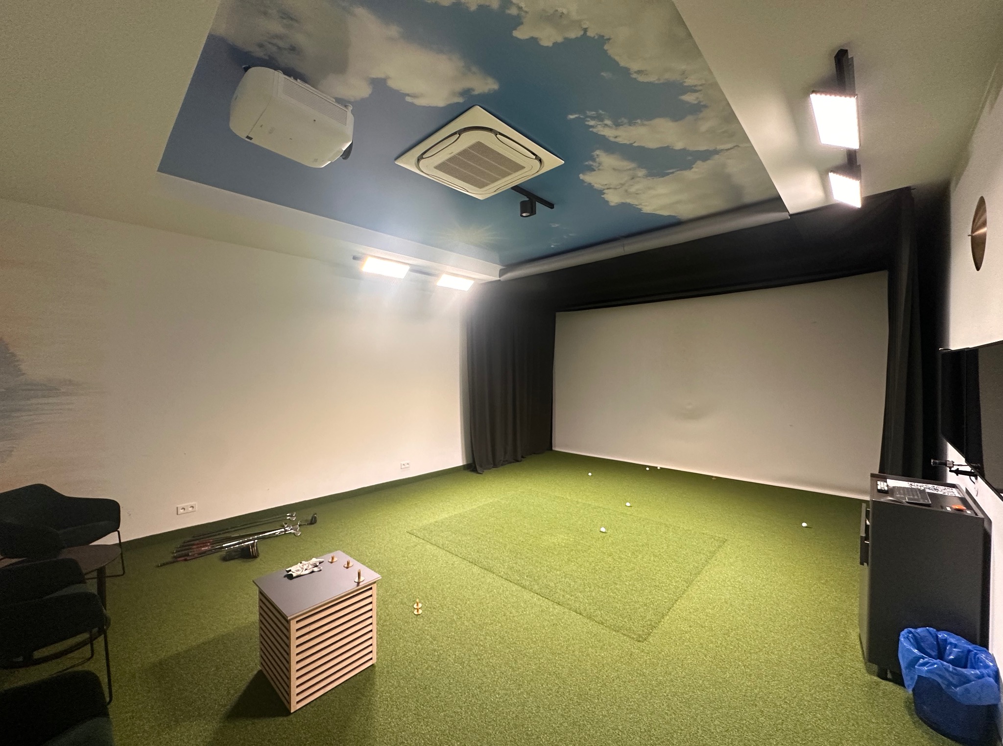 Wnętrze symulatora golfowego: zielona trawa, ekran projekcyjny, sprzęt golfowy i sufit z motywem nieba, tworzące realistyczne środowisko do gry w golfa.