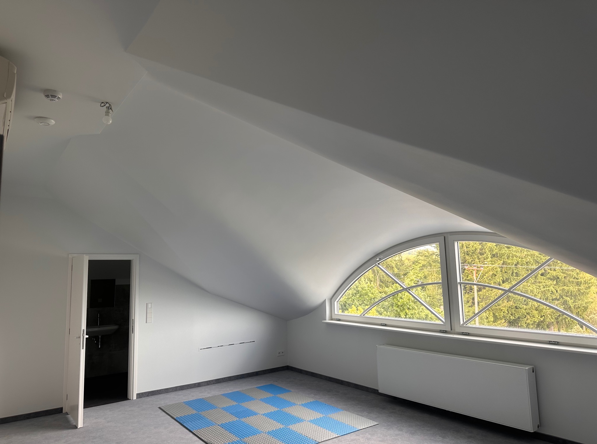 Poddasze po remoncie: białe ściany, okno łukowe z widokiem na zieleń, grzejnik, szara podłoga z niebieską matą. Otwarta biała łazienka. Minimalistyczny styl.