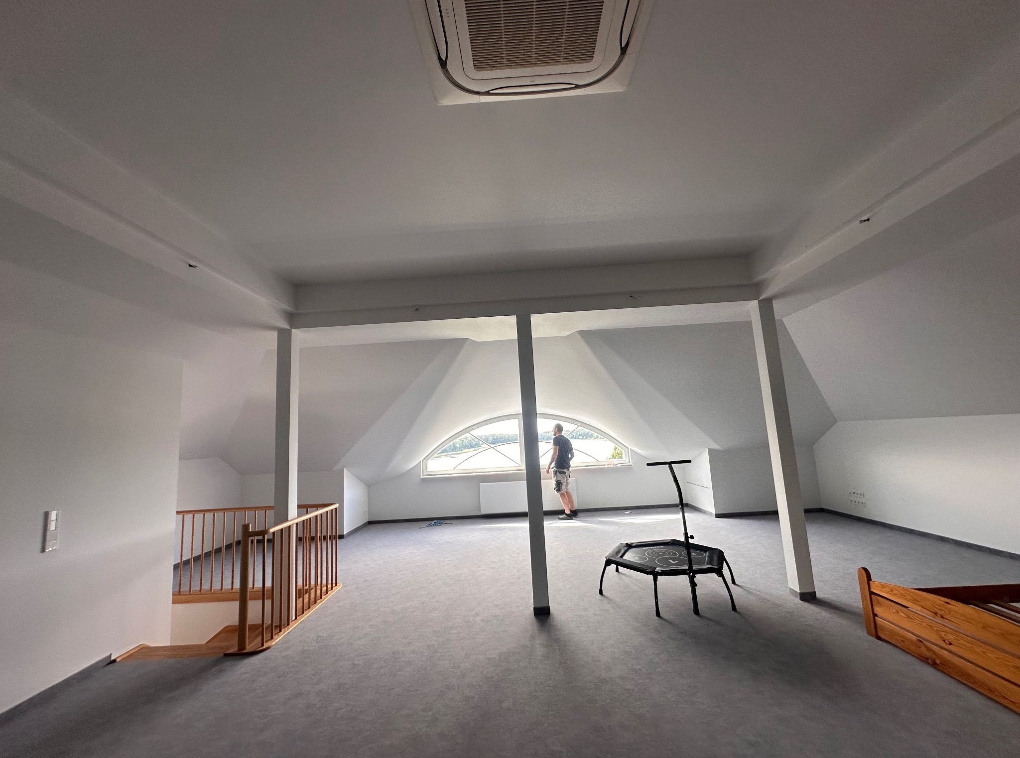 Jasne poddasze po remoncie: gładkie, białe ściany, szara podłoga, okno w kształcie łuku. Mężczyzna przy oknie. Widoczny fragment schodów z drewnianą barierką.