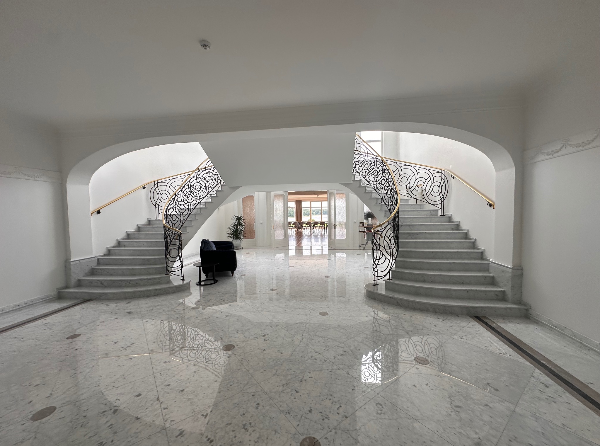 Eleganckie wnętrze holu z marmurową posadzką i symetrycznymi schodami z kutymi balustradami. W tle widoczne przeszklone drzwi prowadzące do dalszej części domu.