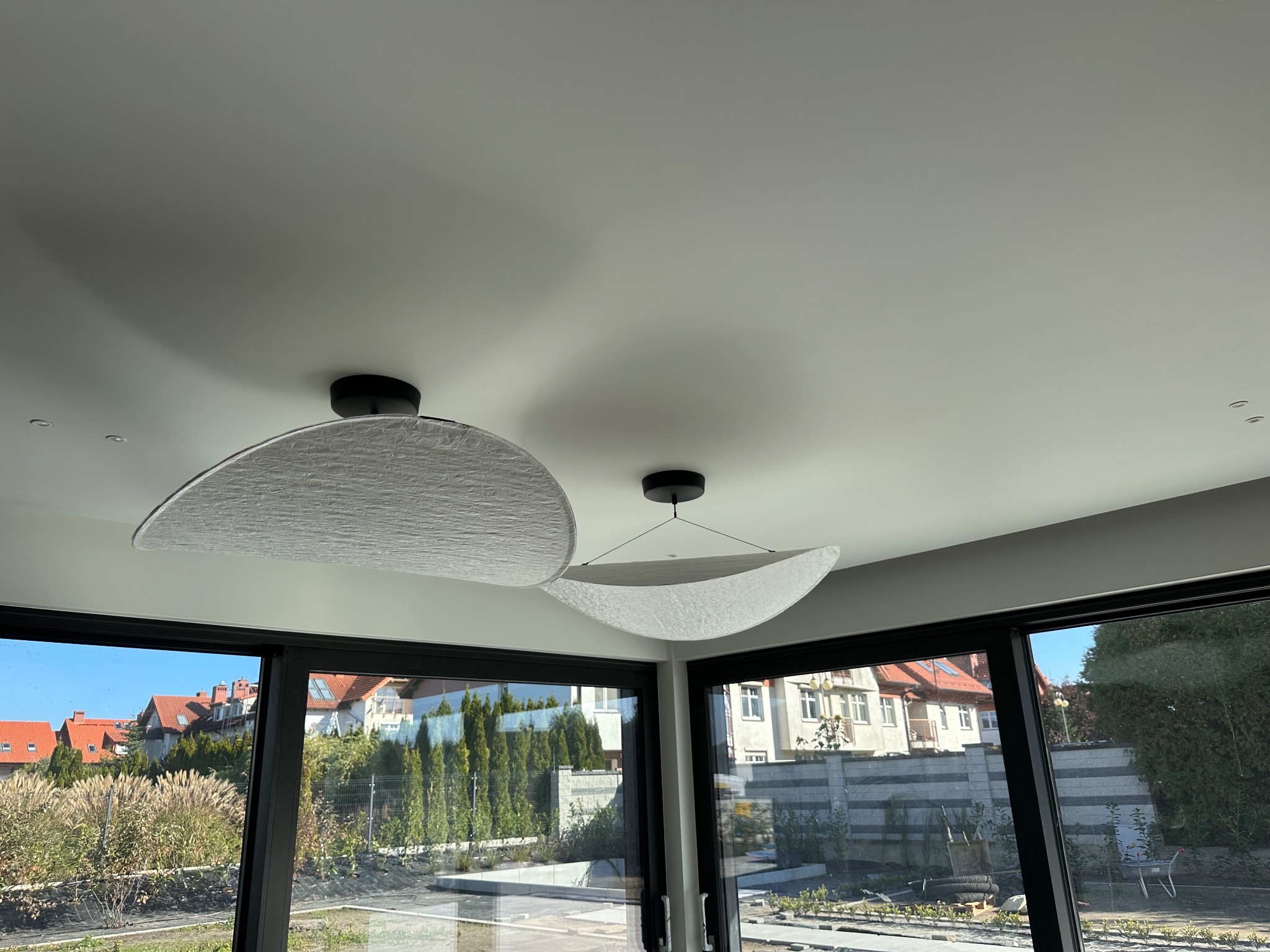 Nowoczesne lampy sufitowe w kształcie półksiężyca, wiszące w jasnym wnętrzu z dużymi oknami, przez które widać zabudowę i zieleń. Minimalistyczny design.