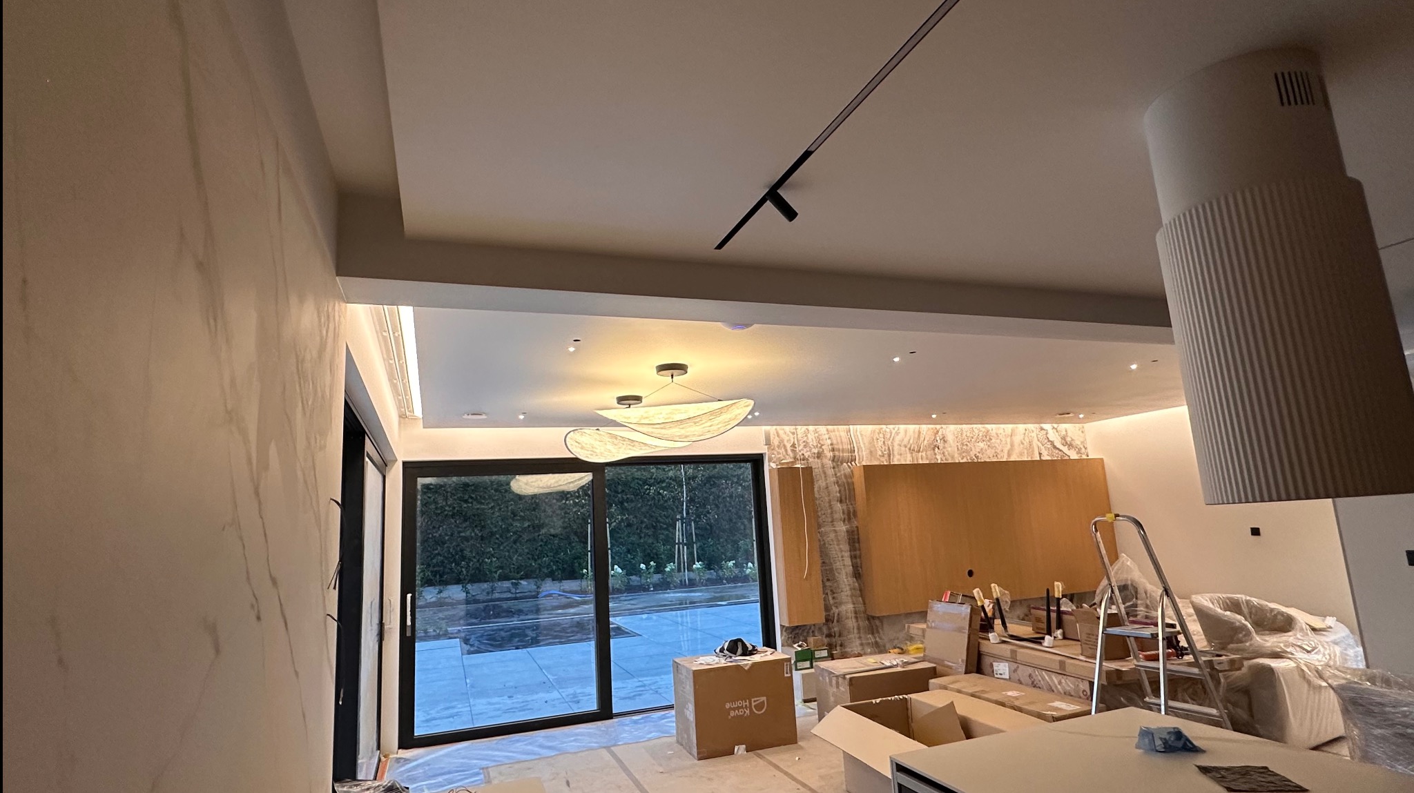 Wnętrze w trakcie remontu: gładkie ściany imitujące marmur, duże okno z widokiem na zewnątrz, kartony na podłodze, drabina oparta o ścianę, nowoczesne oświetlenie.