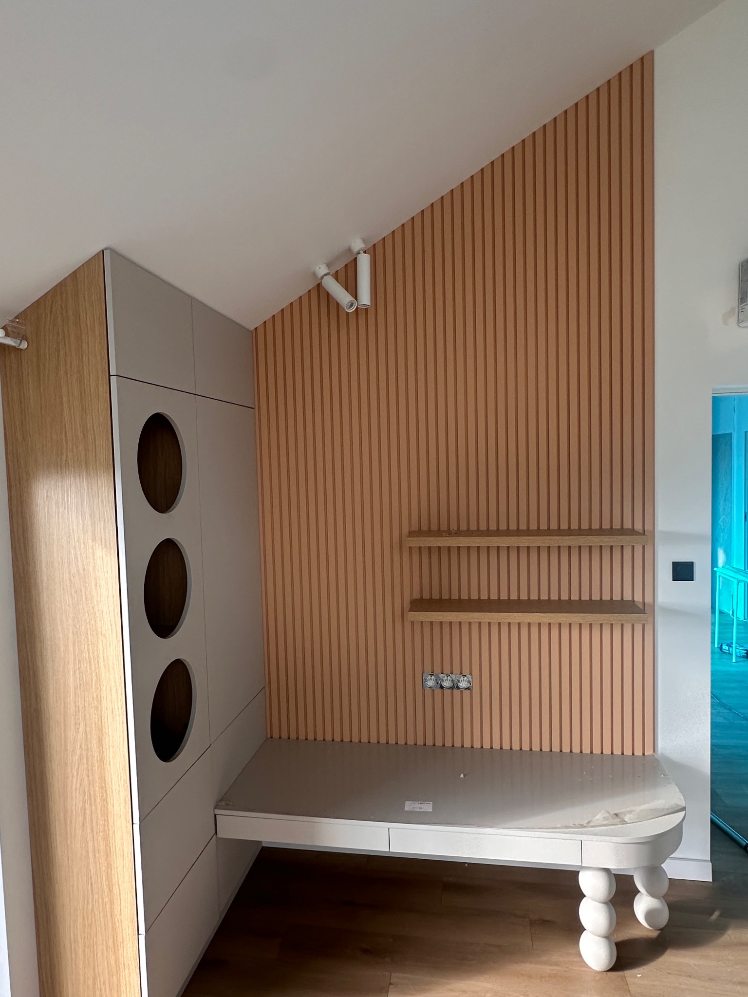 Nowoczesny pokój z zabudową meblową: szafa z okrągłymi otworami, biurko z nogami w kształcie kul i dekoracyjna ściana z drewnianych listew. Oświetlenie punktowe.