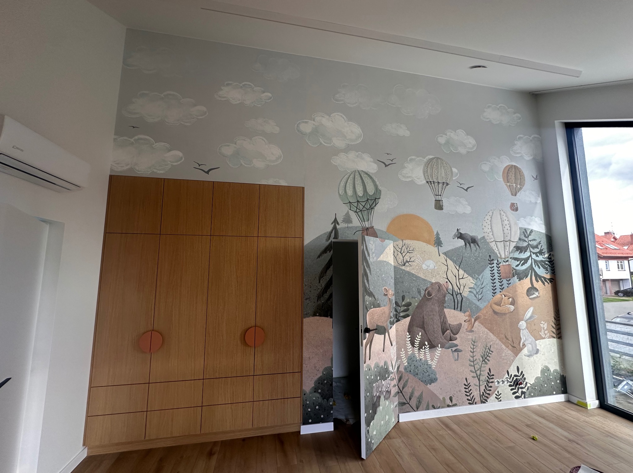 Pokój dziecięcy po malowaniu: ściana z motywem lasu i zwierząt, obok drewniana szafa z okrągłymi uchwytami. Widoczne okno i drewniana podłoga.