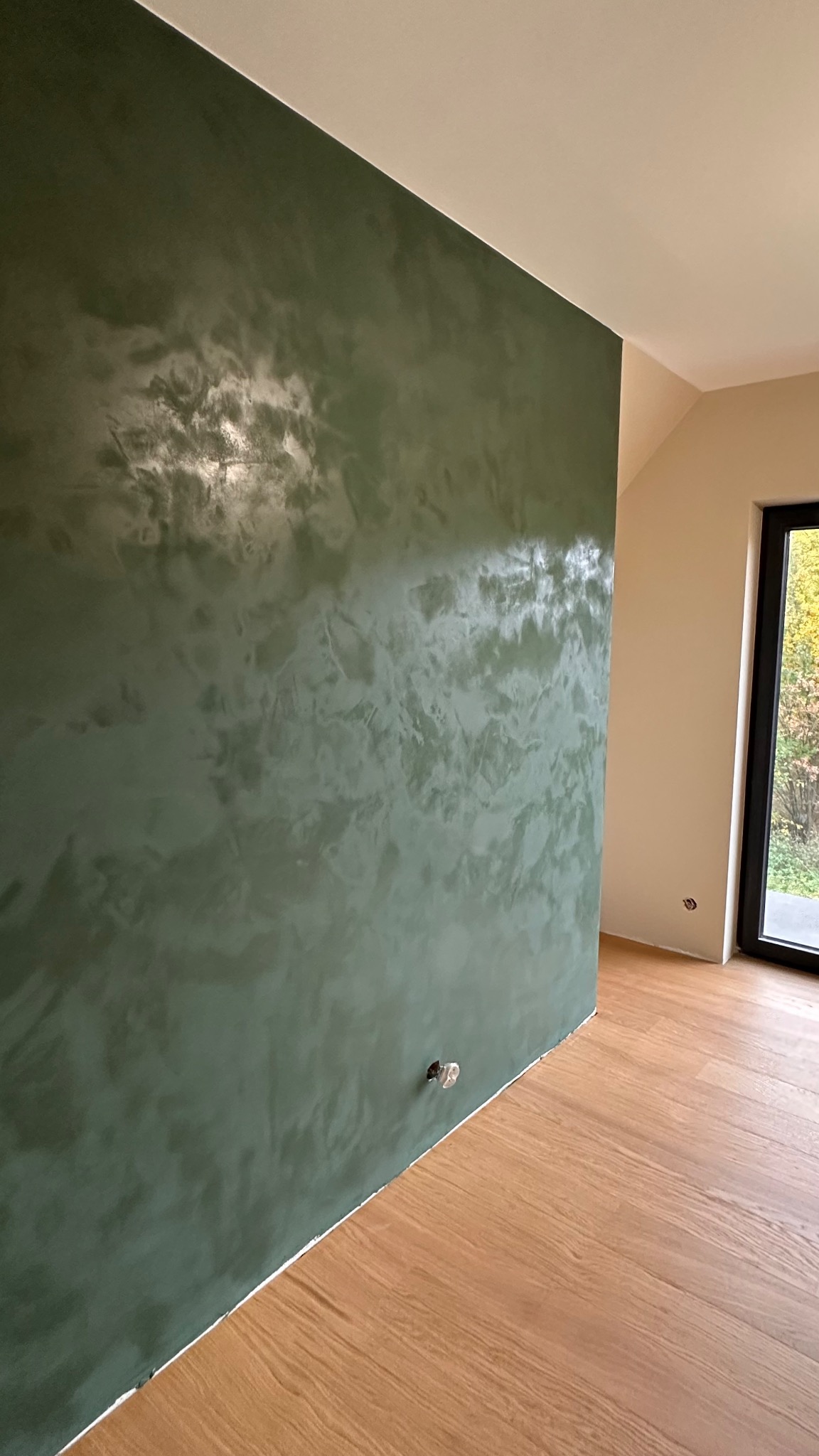 Ściana z dekoracyjną, zieloną fakturą, widoczna pod kątem, z fragmentem okna i drewnianą podłogą. Widoczne gniazdko elektryczne w ścianie.
