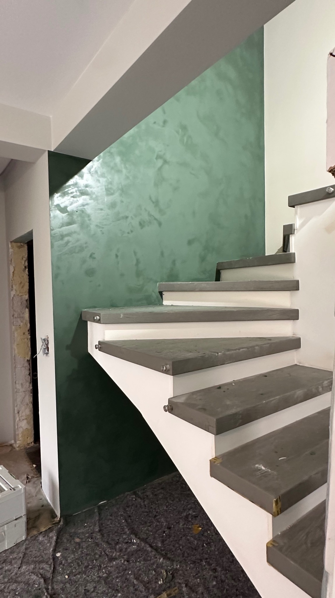 Ściana z efektem dekoracyjnym w odcieniach zieleni sąsiaduje z białymi schodami z szarymi stopniami. Widoczny fragment remontowanego pomieszczenia.